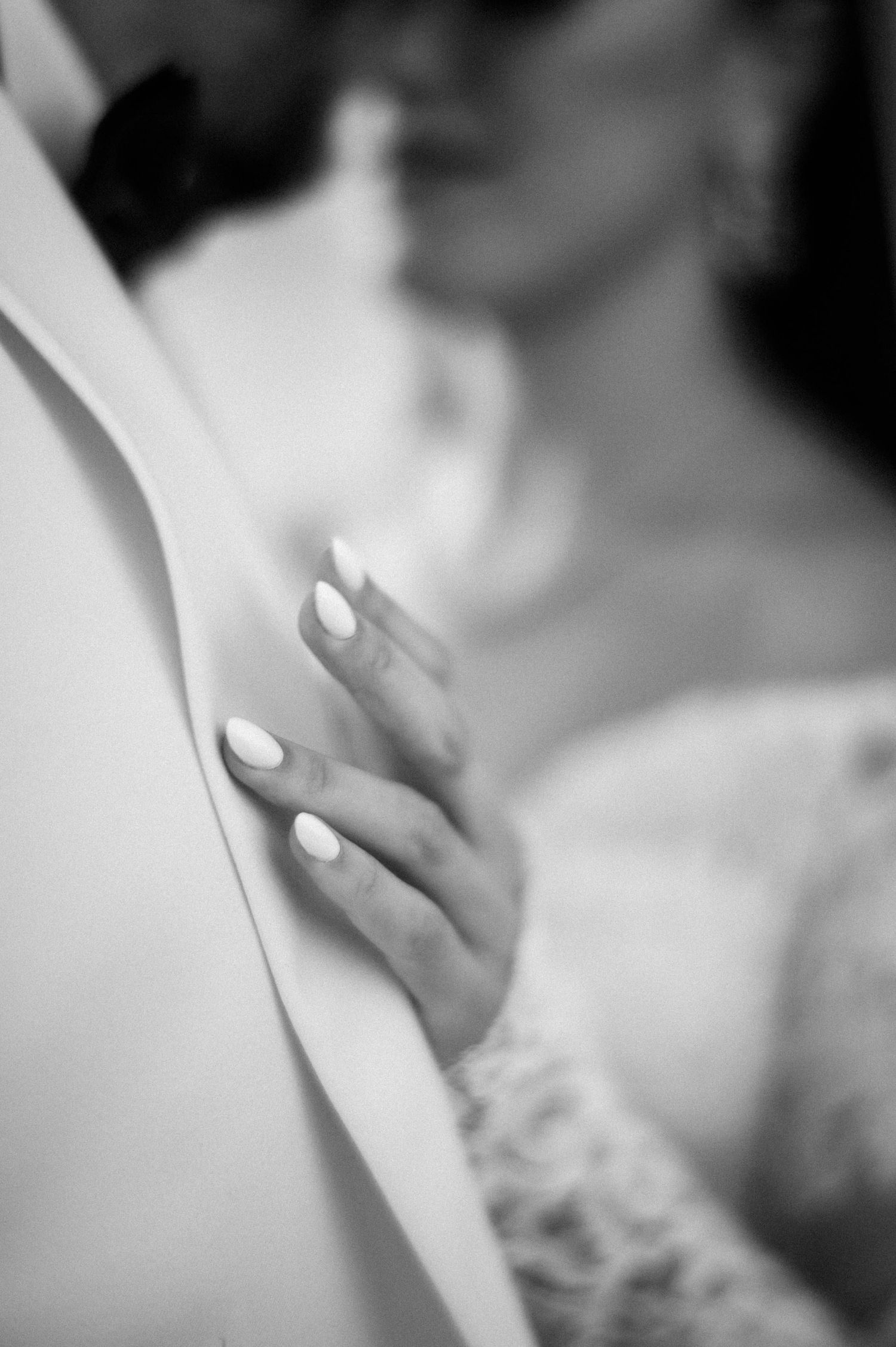 A close-up black and white photo of elegant white manicured fingernails against lace wedding dress fabric.