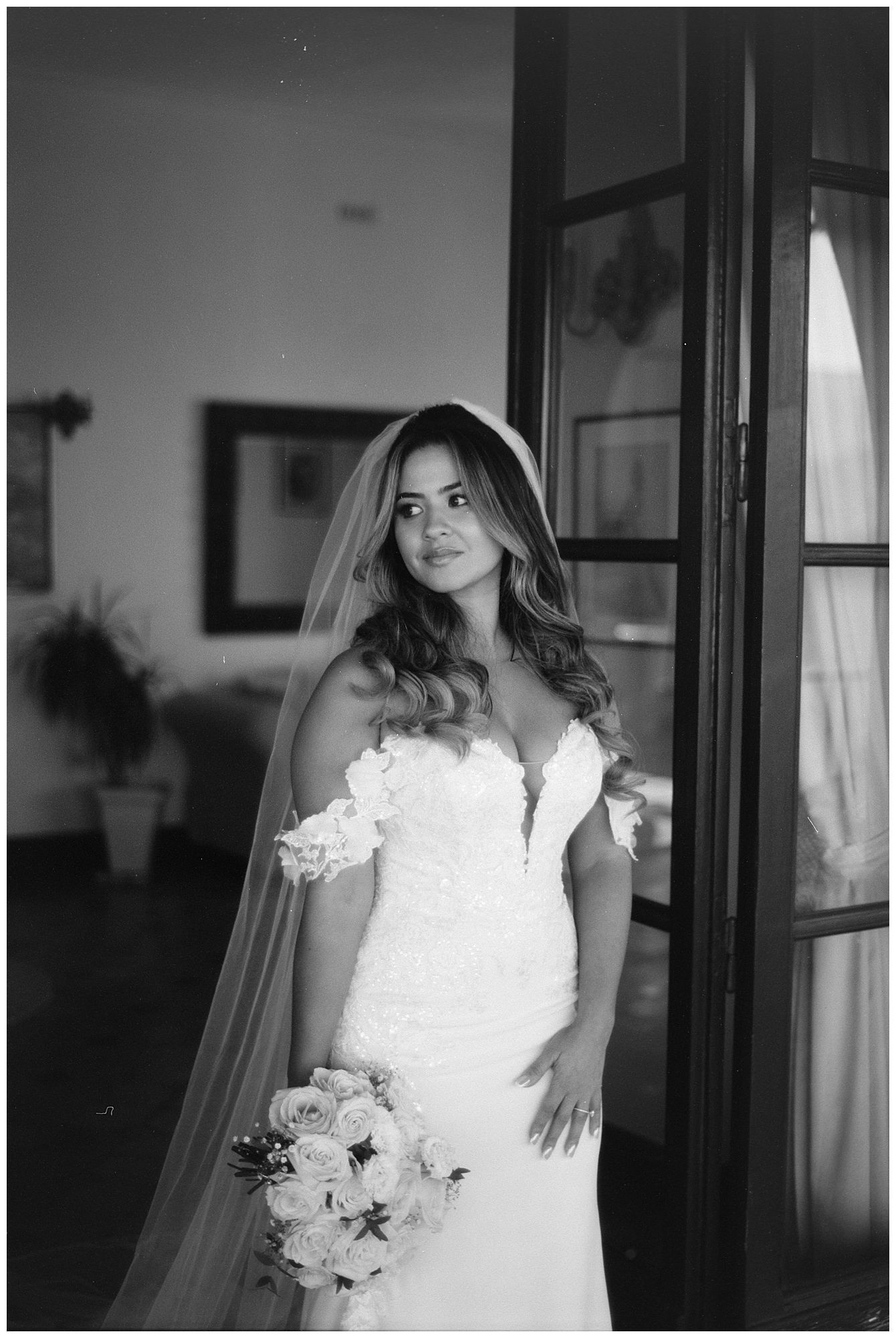 Elegant bride in off-shoulder white lace wedding dress holds bouquet while posing by window in black and white photo.