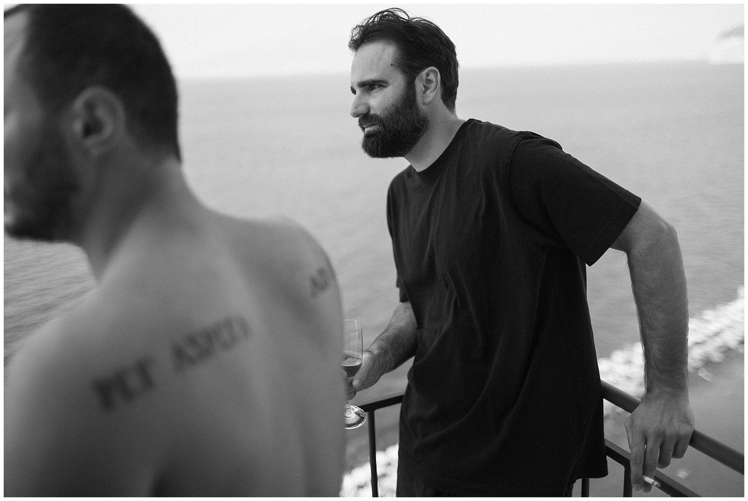 Black and white photograph of two men on a balcony overlooking the ocean, with one wearing a dark t-shirt.