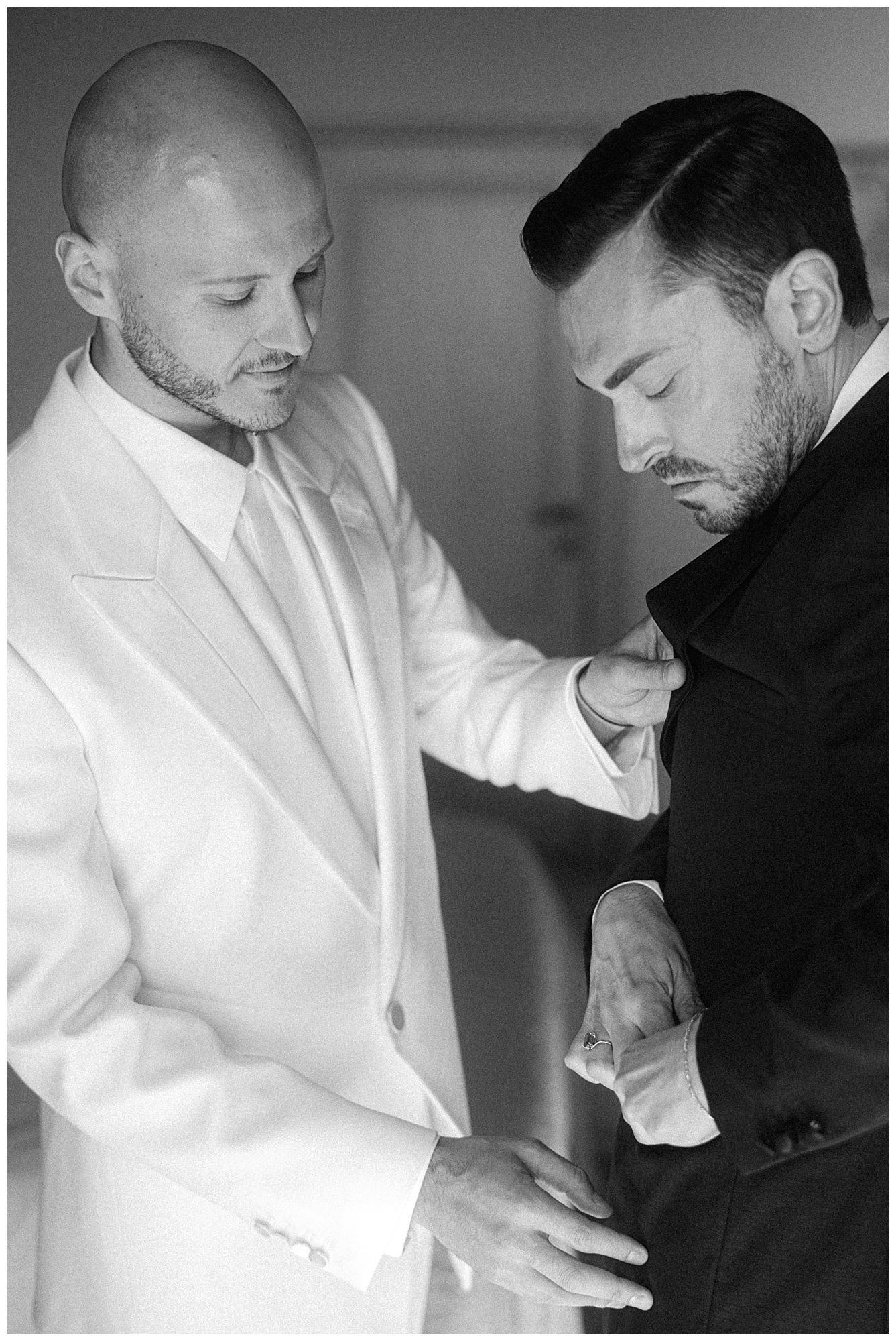 A black and white photograph shows two men, one in a white suit helping adjust the clothing of another in black attire.