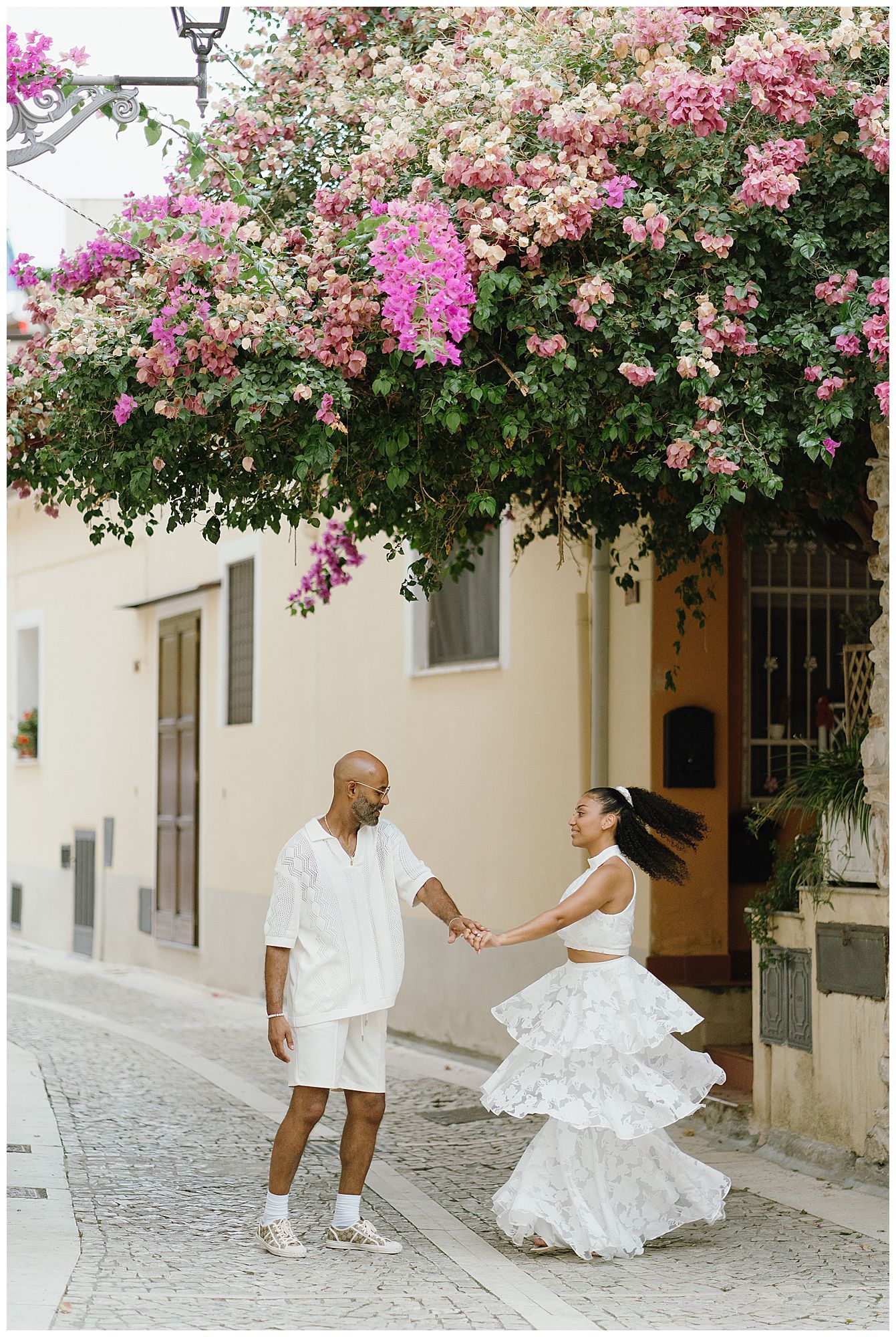 A couple holds hands beneath a blooming bougainvillea arch on a quaint cobblestone street in Mexico.