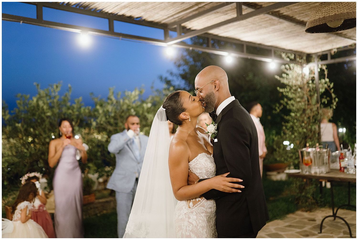 A romantic outdoor wedding celebration showing couples dancing and embracing under twinkling lights at dusk.