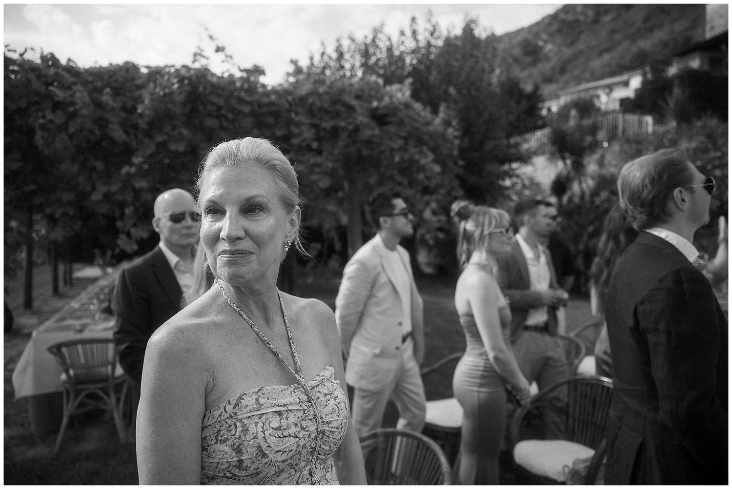 Black and white outdoor social gathering with guests mingling on a terrace with mountain scenery in the background.