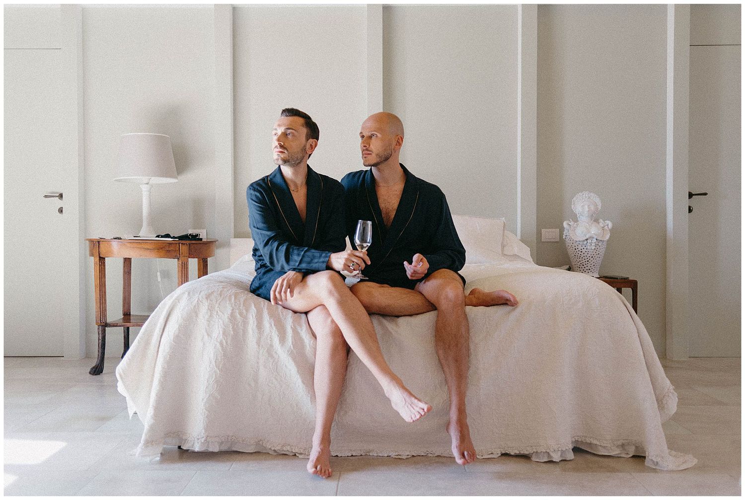 Two people in dark clothing sit casually at the edge of a white bed in a minimalist bright room.