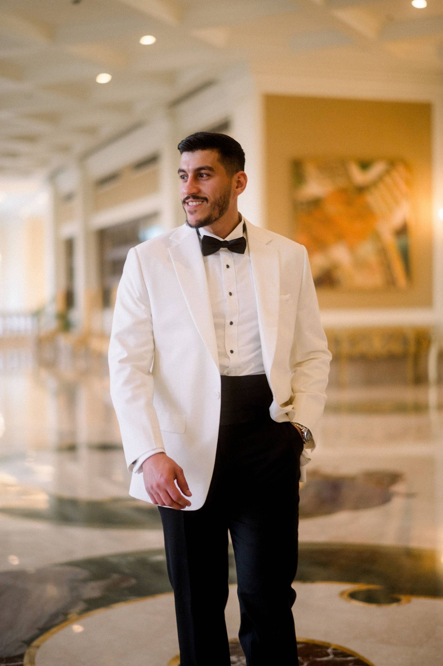 Someone wearing a classic white dinner jacket with black bow tie and trousers stands in an elegant hallway.