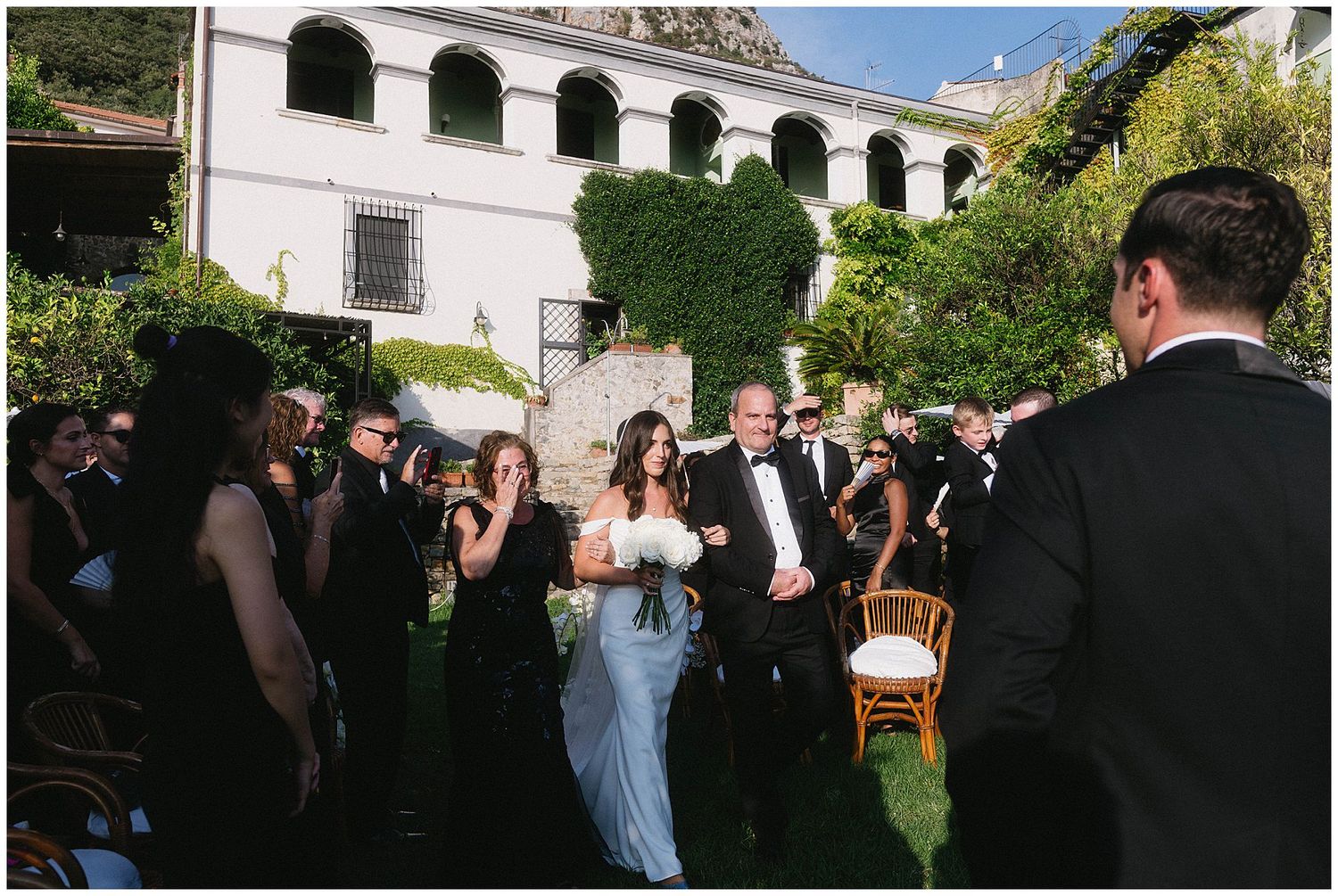 Wedding party gathered in an outdoor courtyard with ivy-covered white walls and arched architecture in the background.