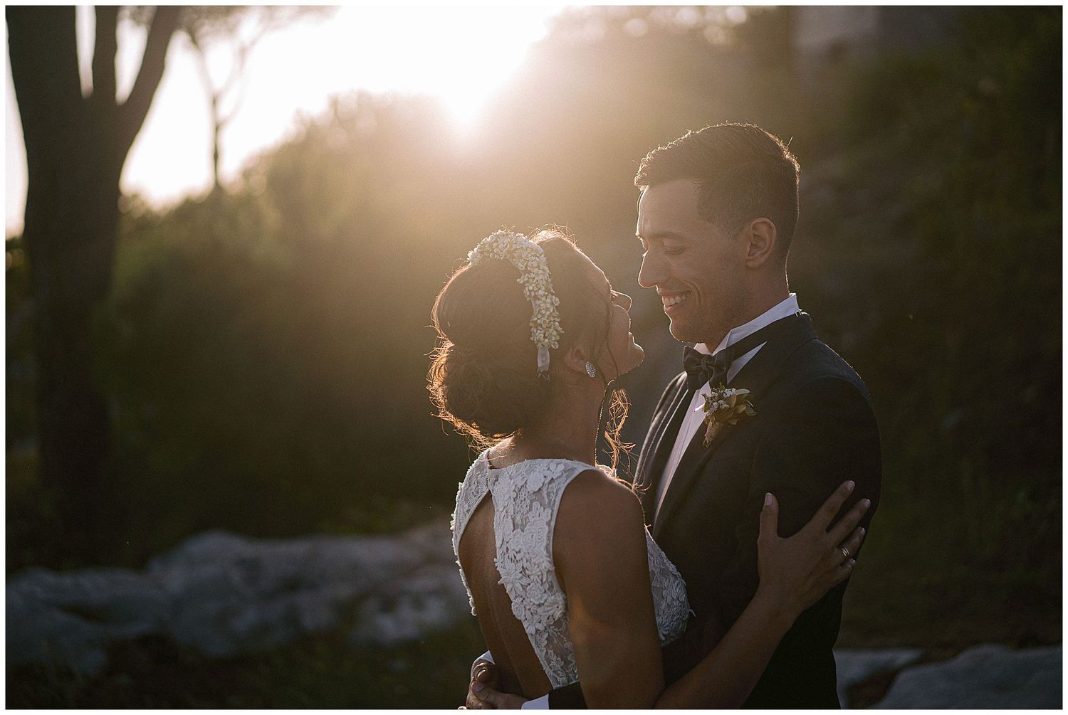 Couple embracing in elegant wedding attire during golden hour sunset with backlit silhouettes and romantic atmosphere.