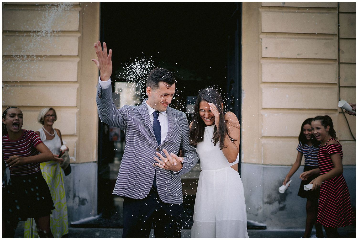 Newlyweds celebrate with confetti and joyful expressions while exiting a building surrounded by wedding guests.