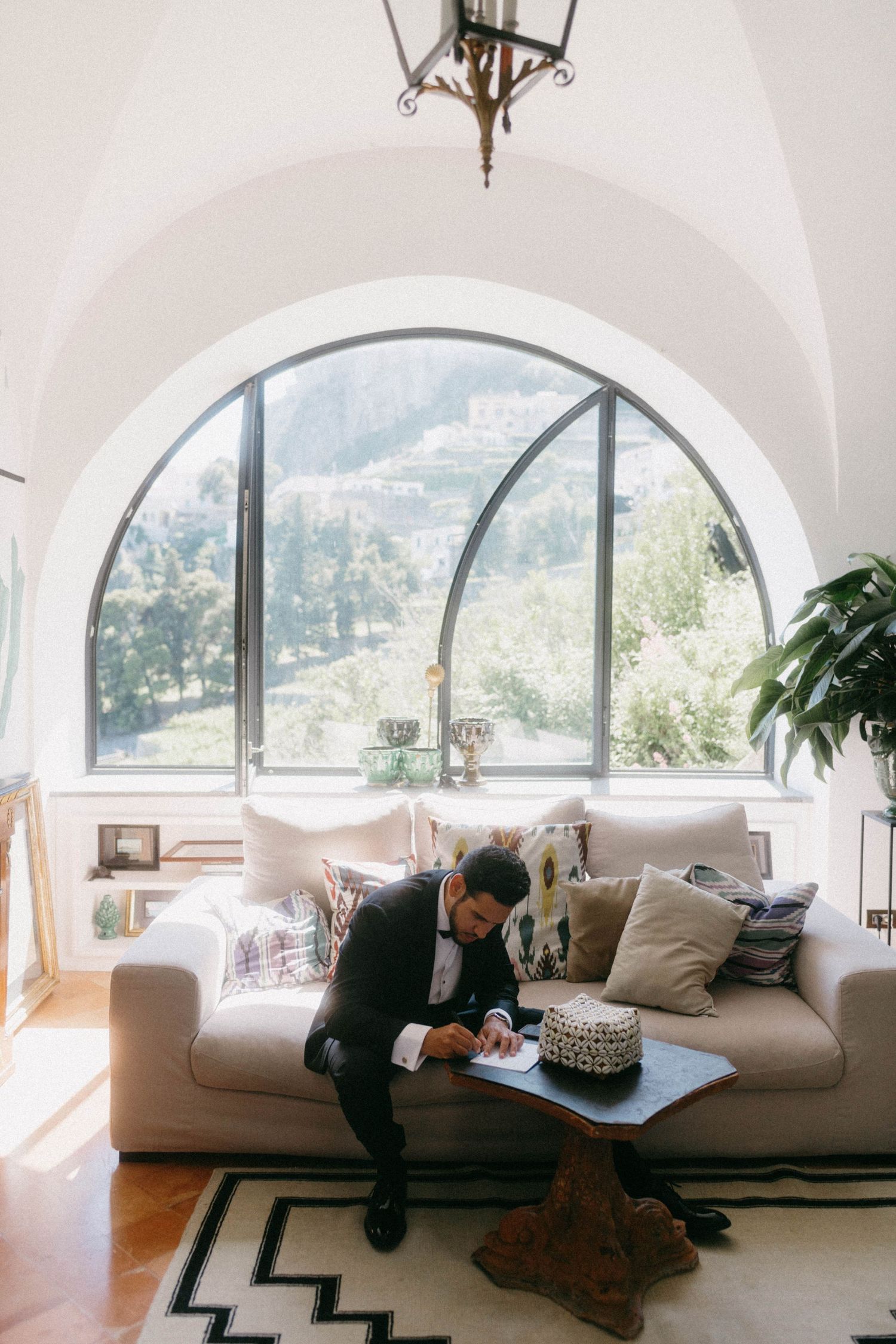 Modern living room with large arched window overlooking mountains and person relaxing on beige sofa.