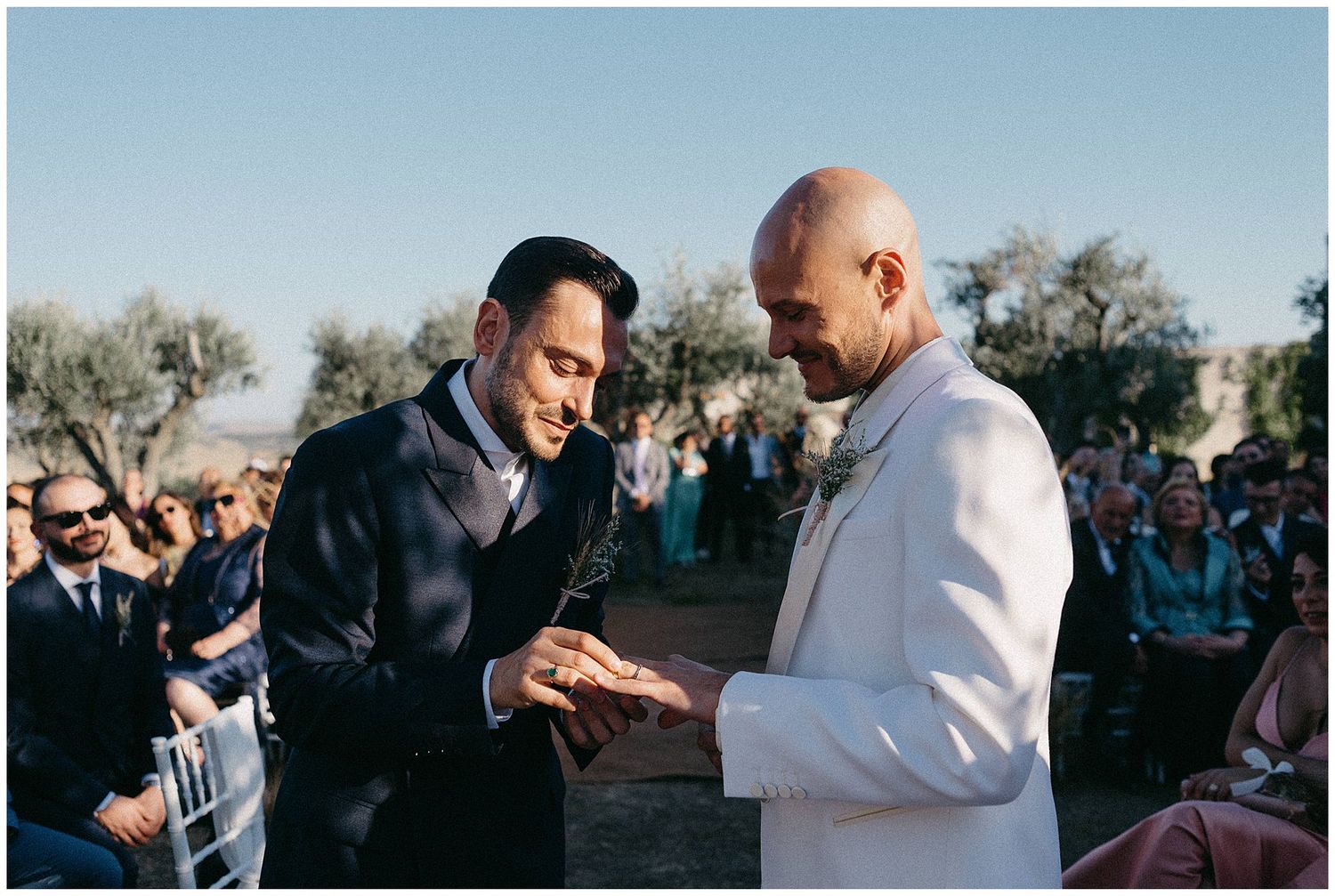 A touching moment during an outdoor wedding ceremony shows two people exchanging rings in front of gathered guests.