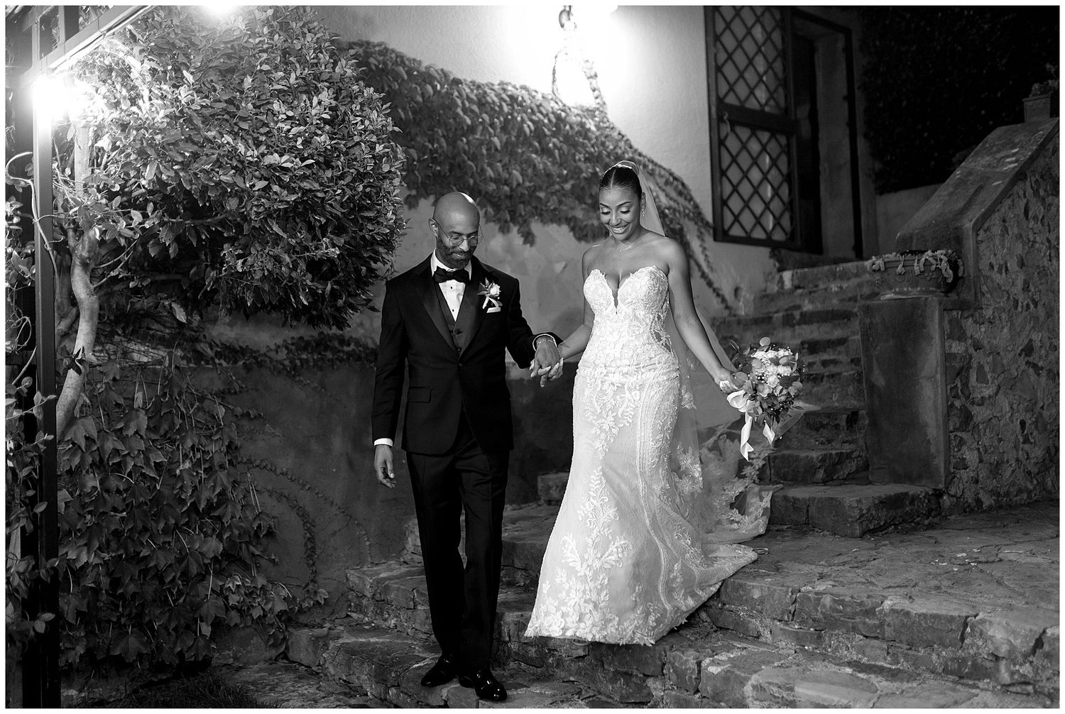 A couple in elegant wedding attire descends stone steps at a romantic outdoor venue in black and white.