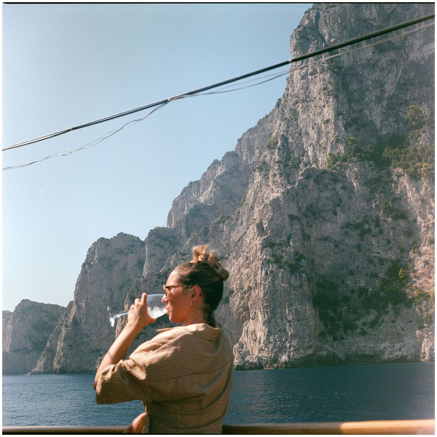 Person wearing sunglasses drinks from a bottle while aboard a boat near steep coastal cliffs by the sea.