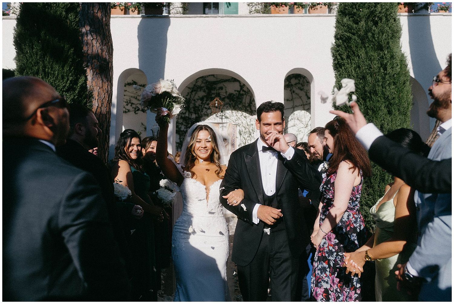 A newlywed couple exits their wedding ceremony while guests surround them at an elegant venue with white arches.