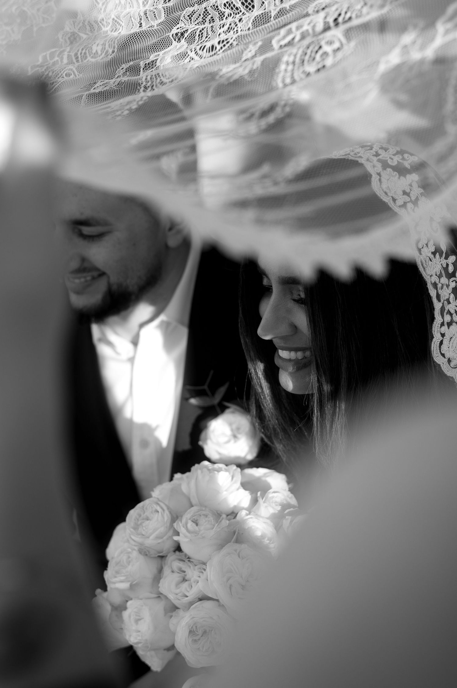 Black and white photo of newlyweds under a veil with a bouquet of white roses.