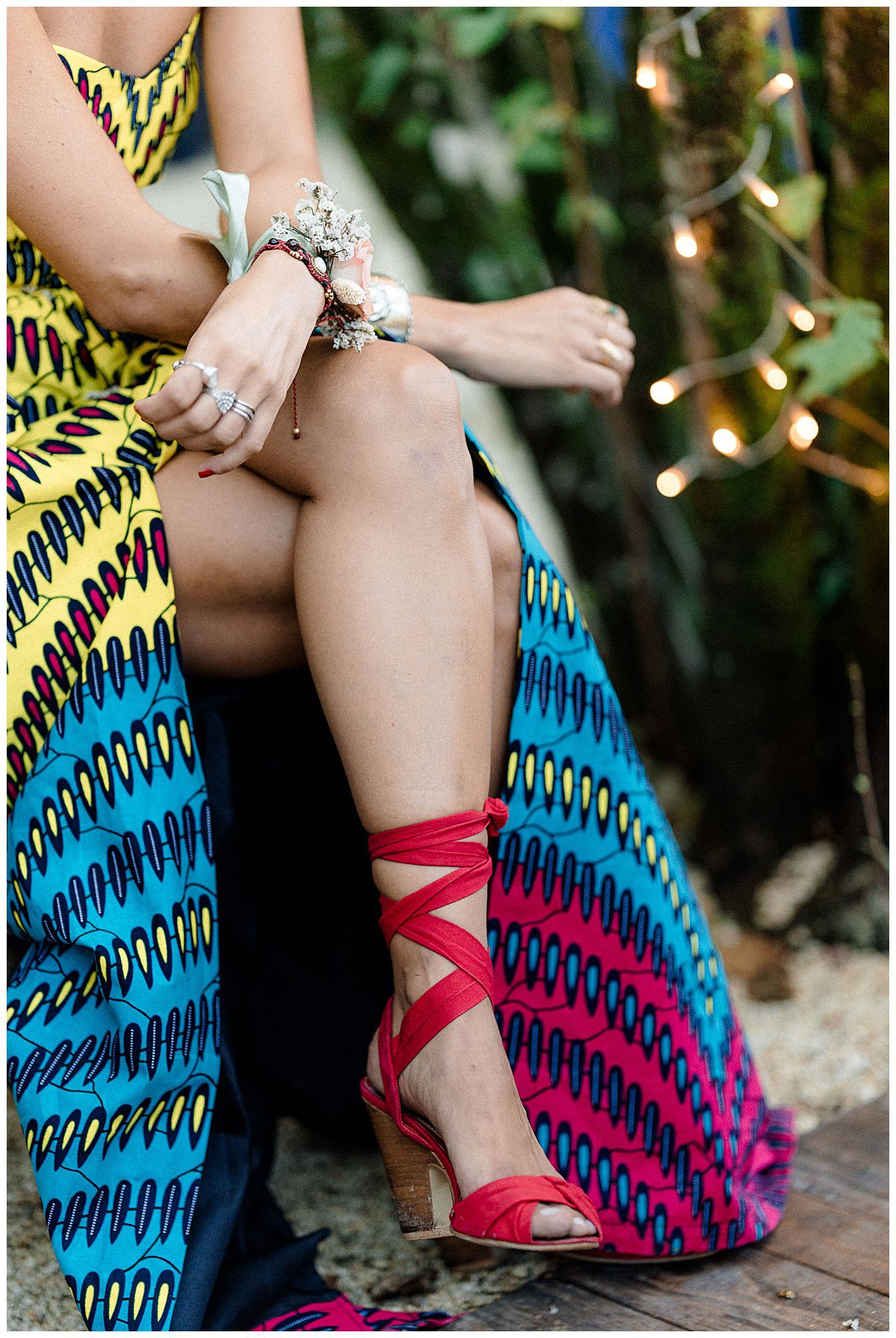 Woman in colorful printed dress wearing red strappy sandals with twinkle lights in background.
