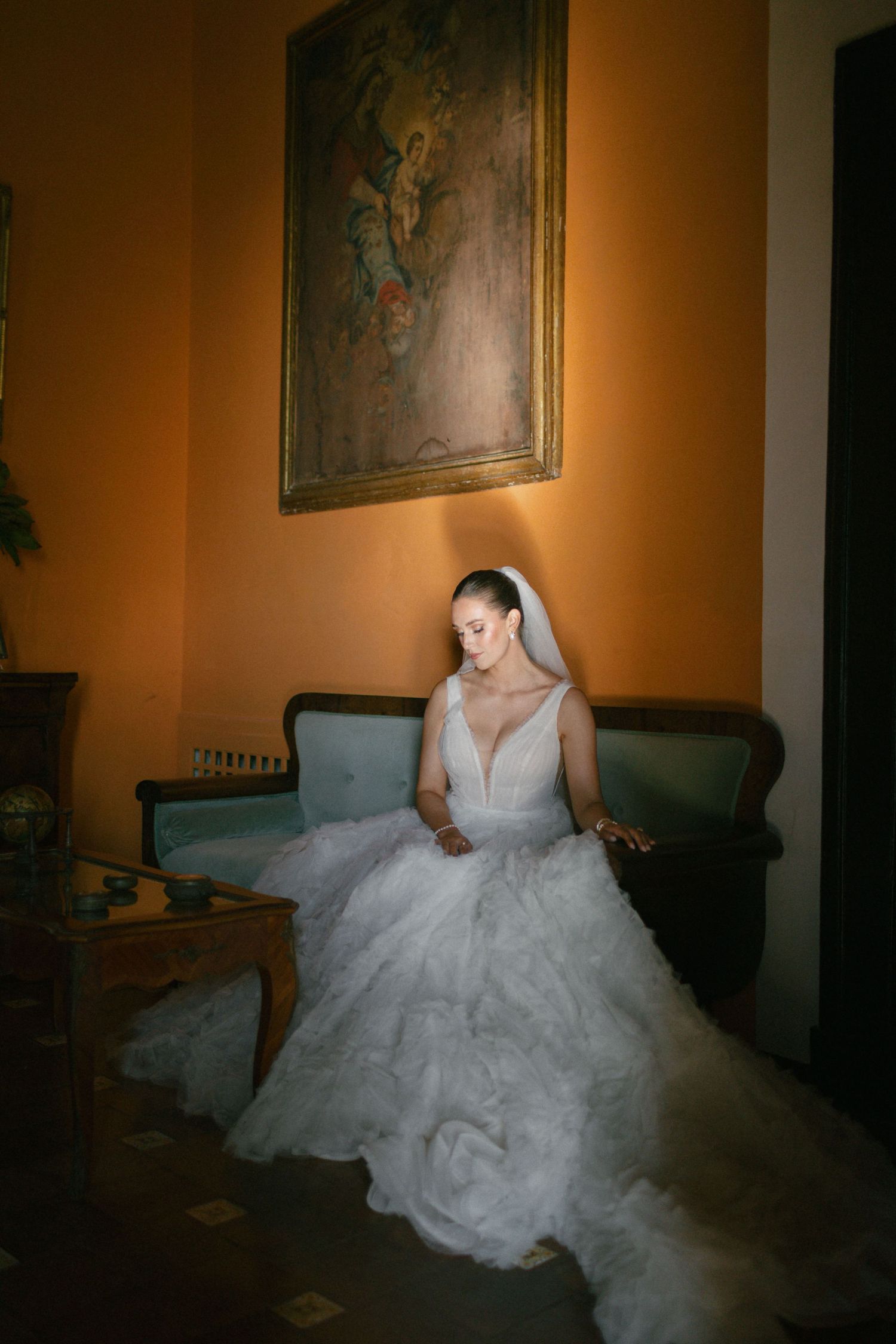 A bride in a flowing white dress sits elegantly on a vintage couch in a warmly lit room with ornate decor.
