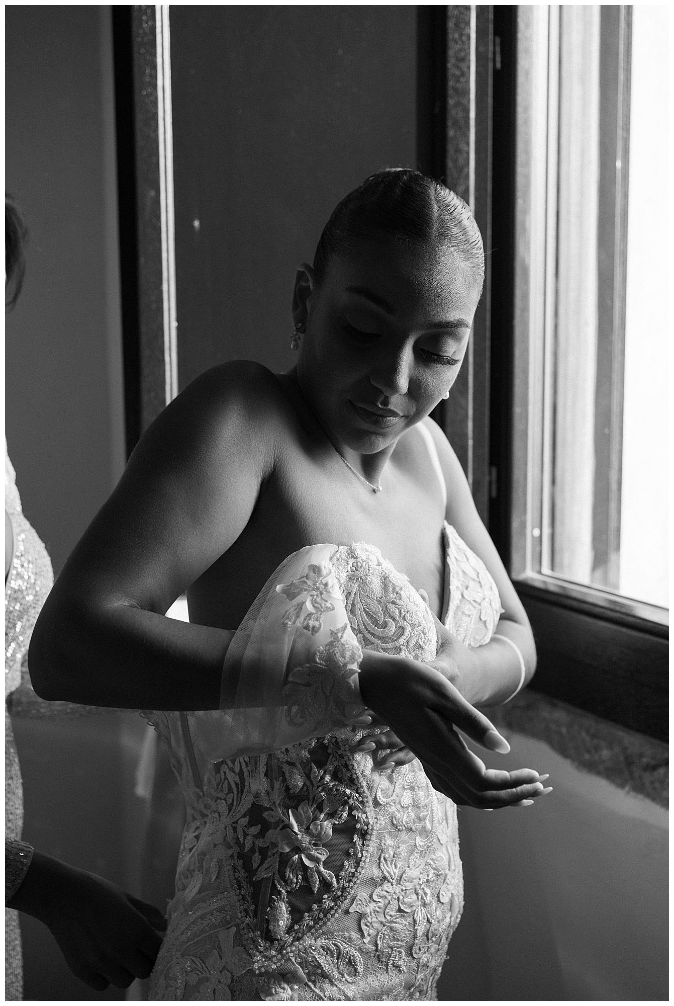 A black and white portrait of someone in a lace wedding gown adjusting their dress by a window.