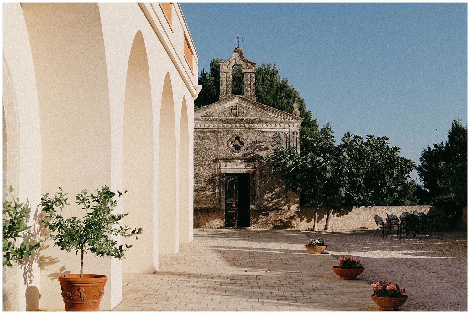 Mediterranean style stone church with arched walkway and potted plants under bright blue sky.