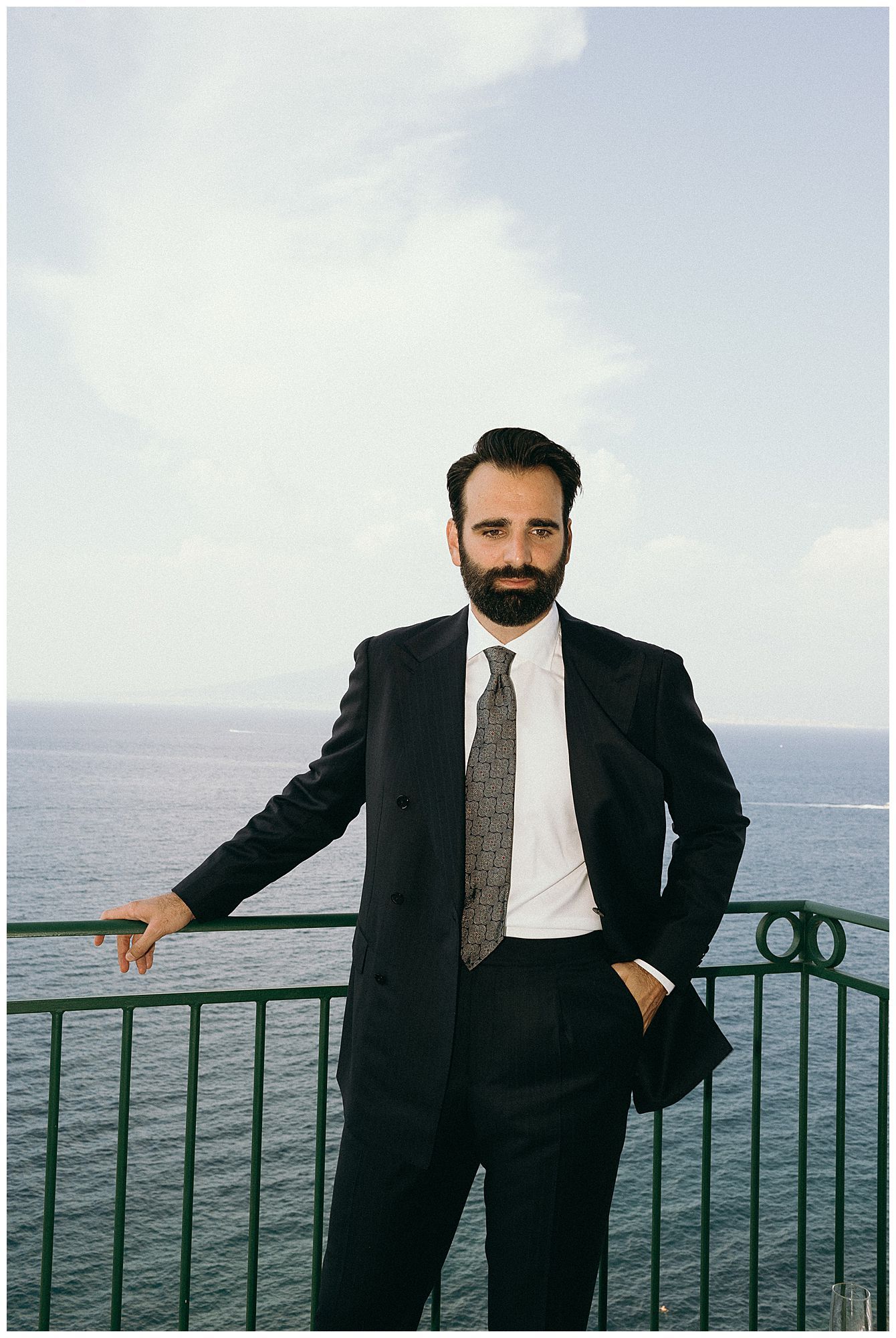 Professional in a dark suit and tie stands at an oceanfront balcony railing with a scenic coastal view in the background.