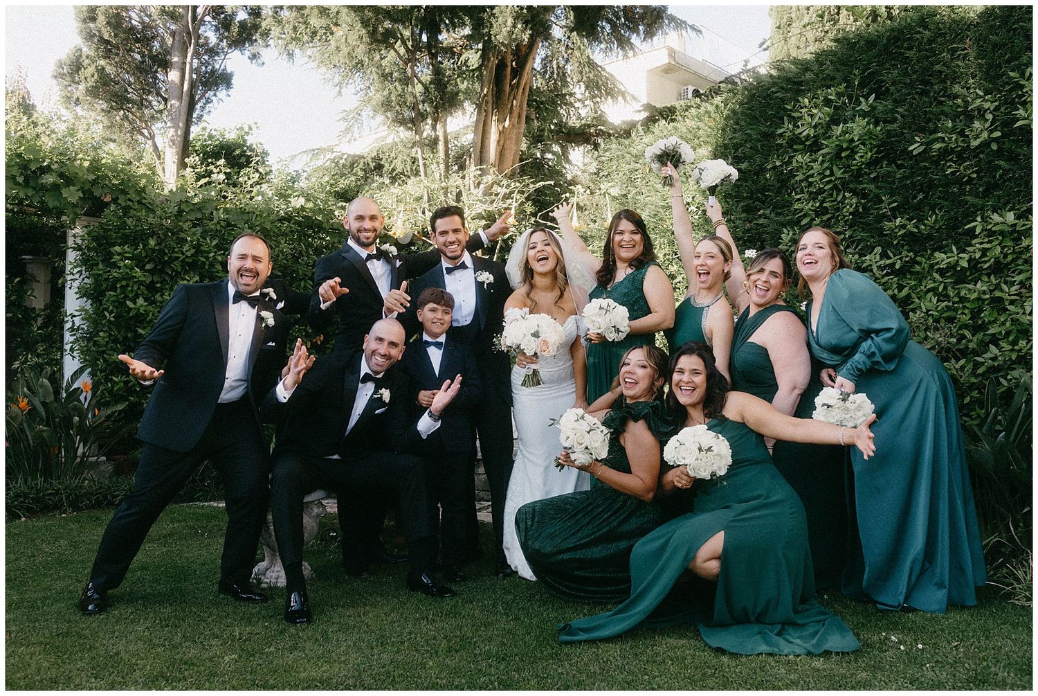 Wedding party in formal attire with bridesmaids in dark green dresses poses joyfully in a garden setting.
