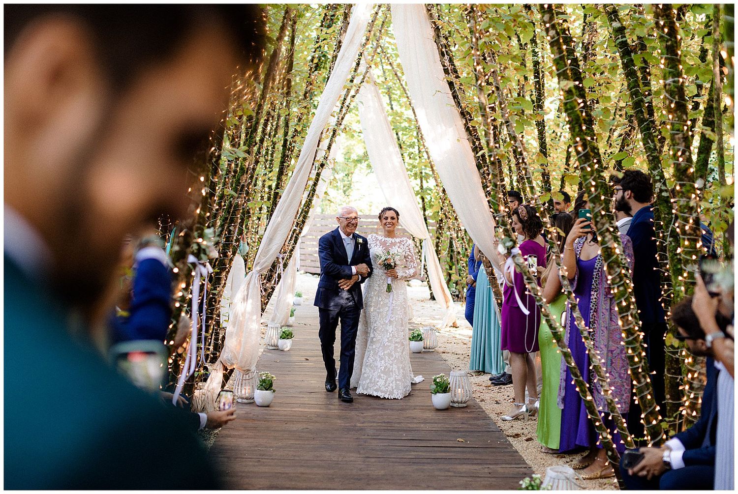 A joyful outdoor wedding ceremony on a wooden walkway decorated with twinkling lights and greenery.