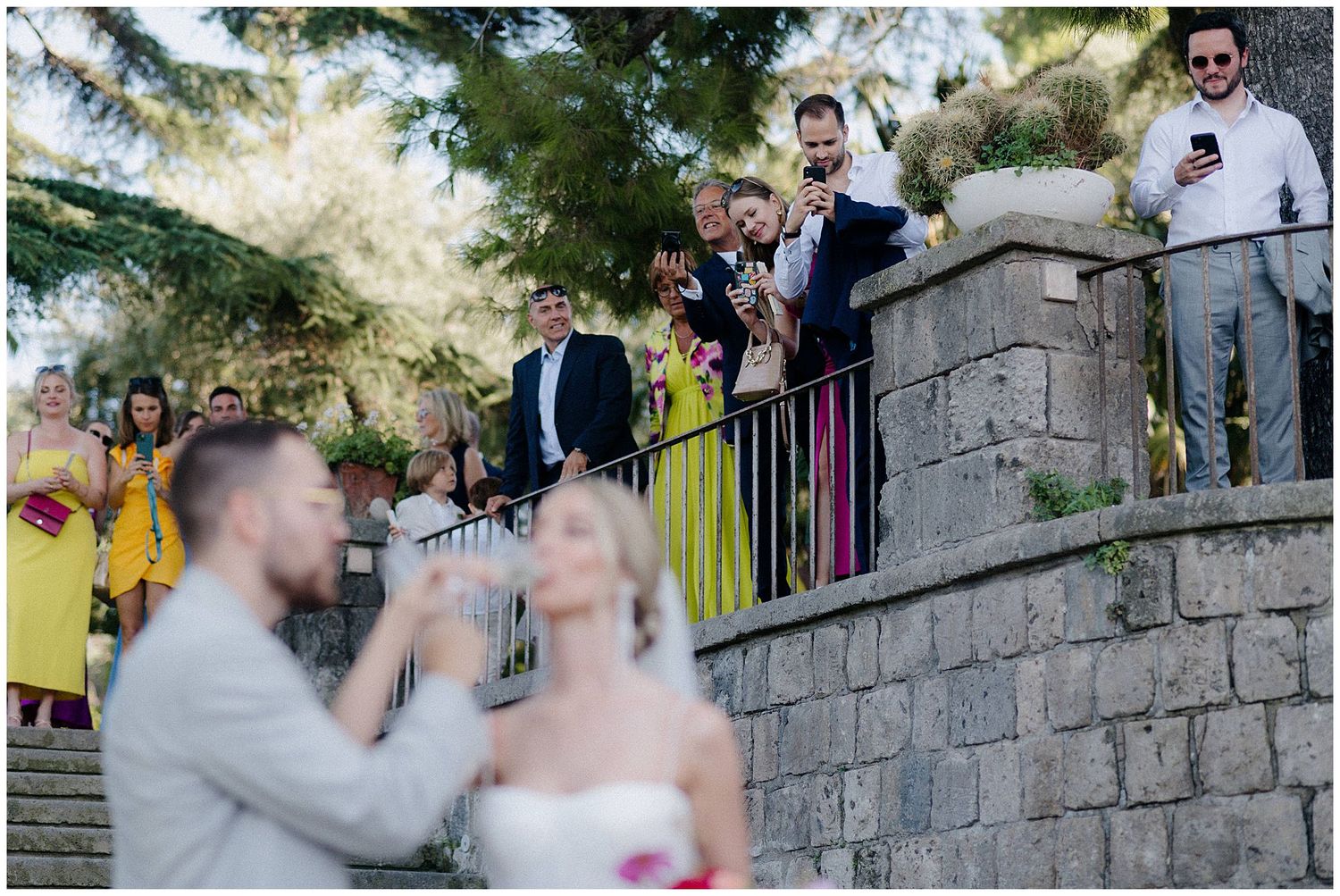 Wedding guests watch from a stone wall as a couple celebrates on the steps below during an outdoor ceremony.