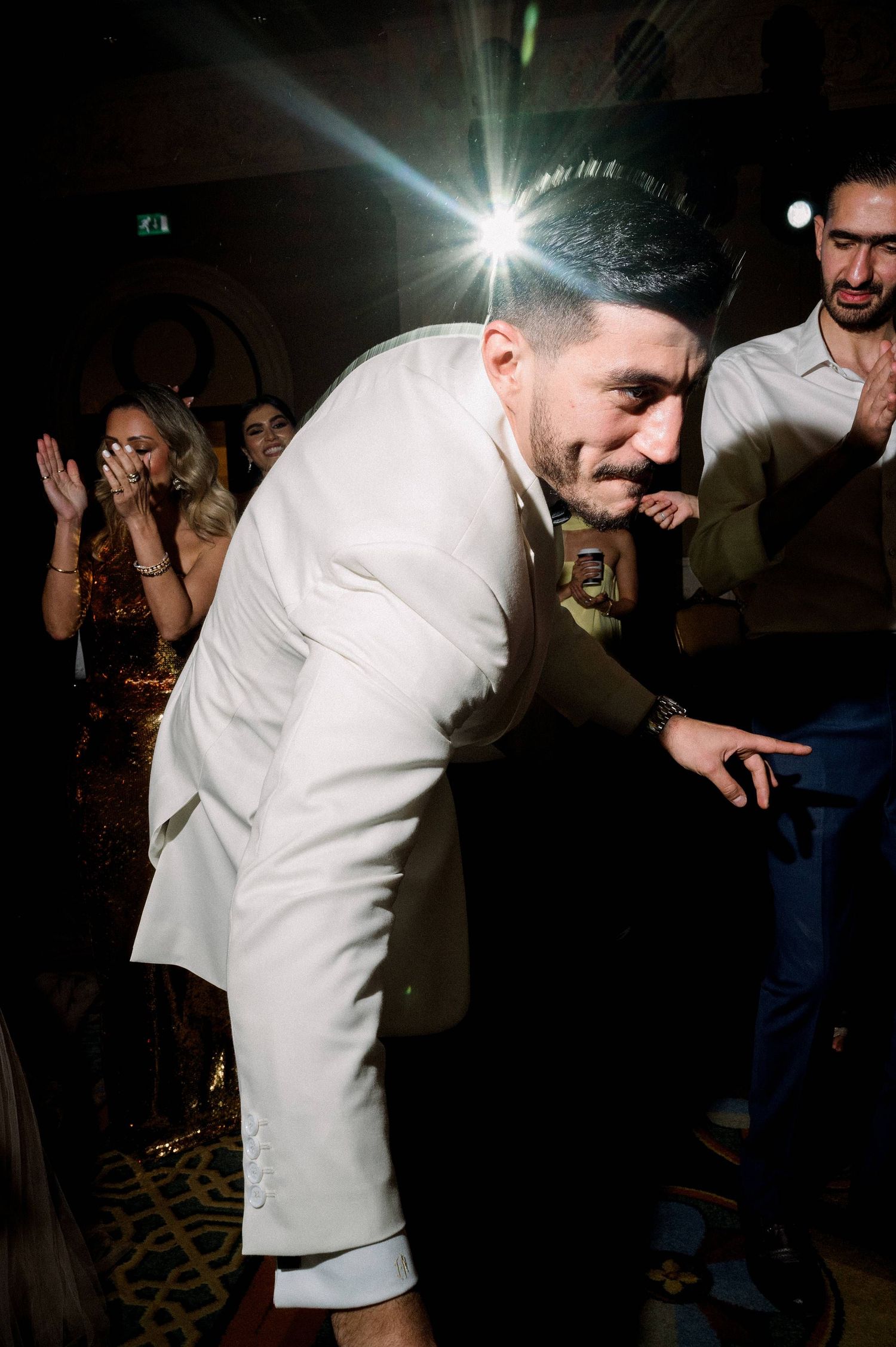 Person in a white suit dancing and leaning forward at a nightclub with bright lights and people in the background.