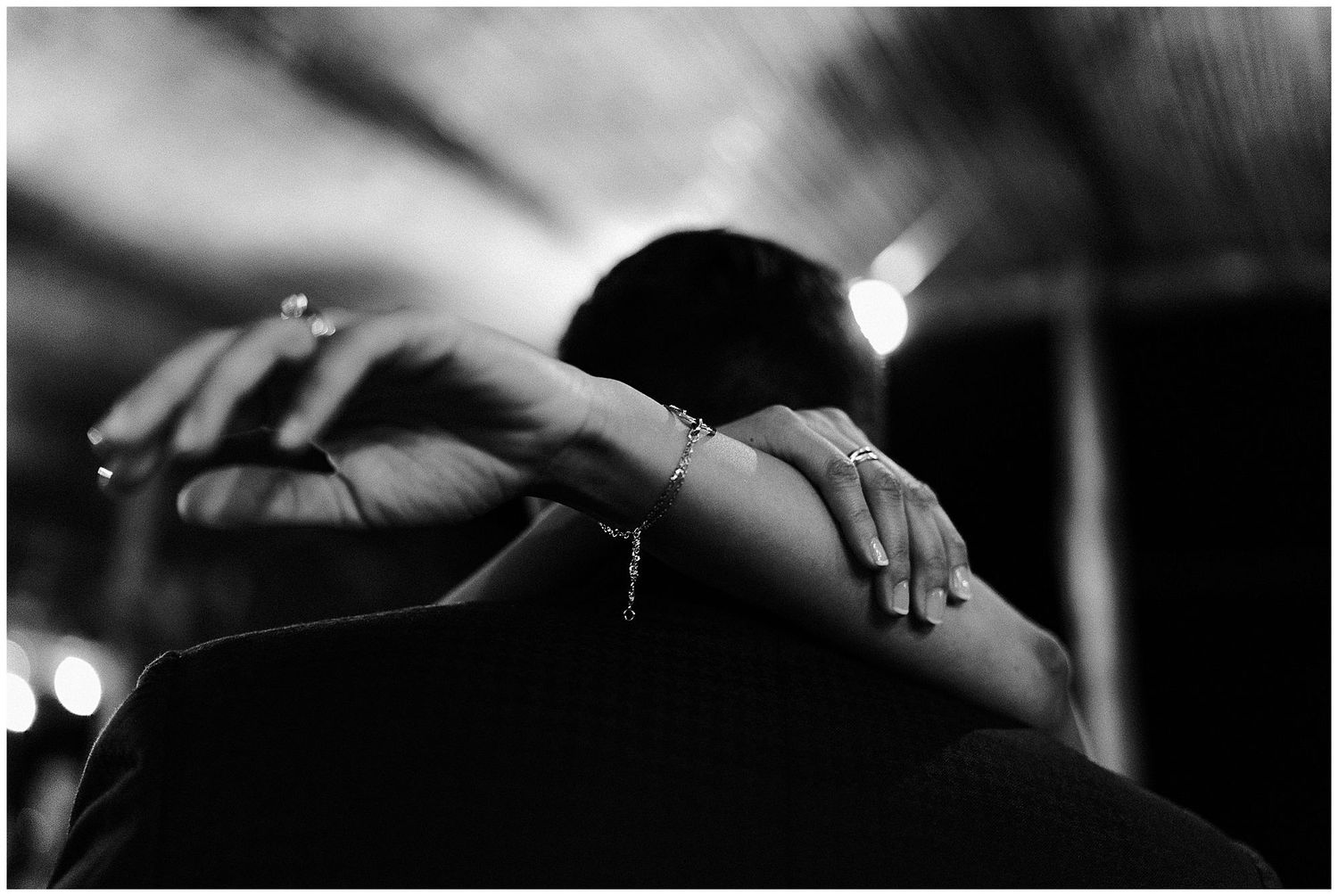 Black and white photograph showing embracing silhouettes with hands and rings visible in soft focus lighting.
