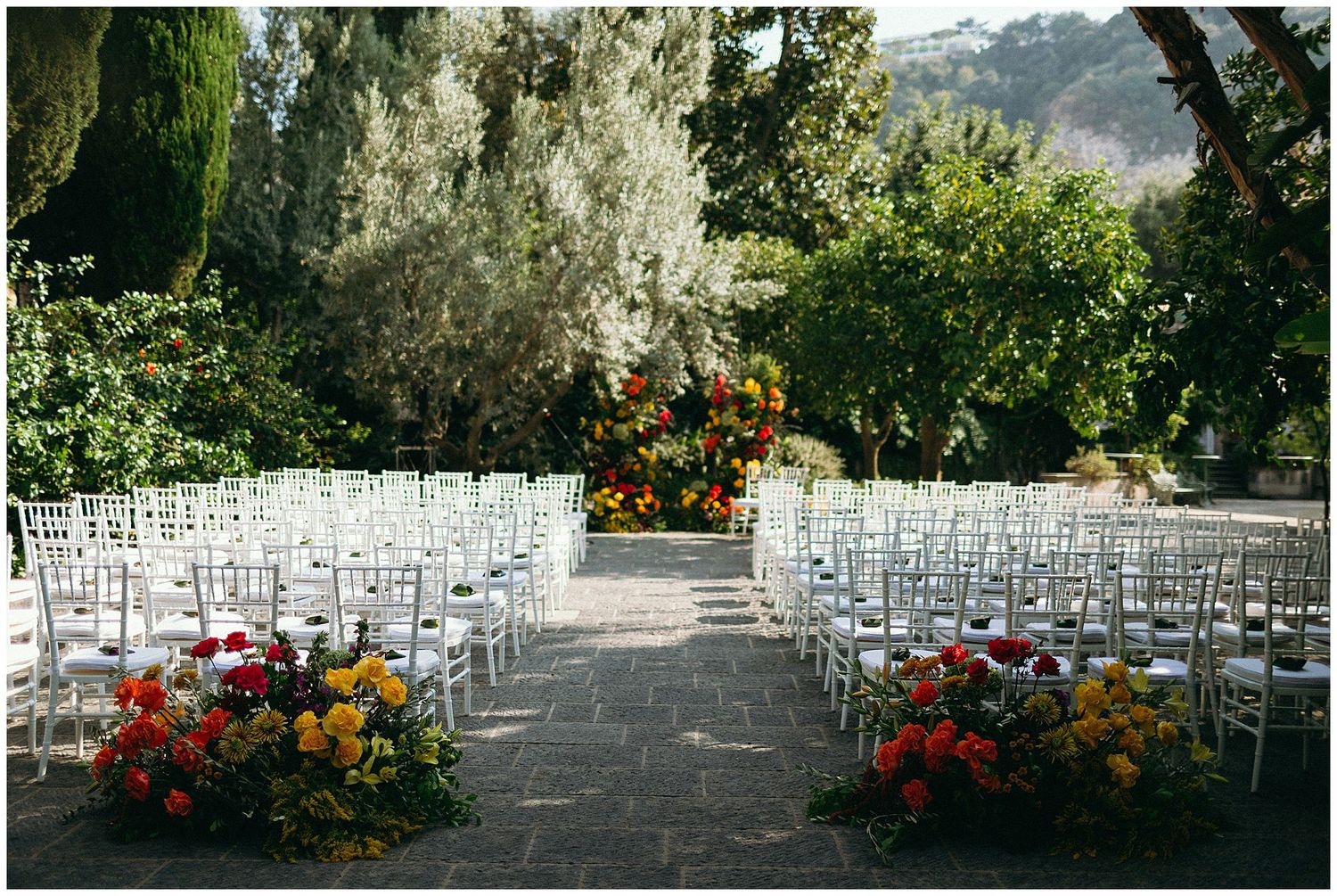 White ceremony chairs set up in rows on stone patio with colorful flower arrangements and greenery for outdoor wedding.