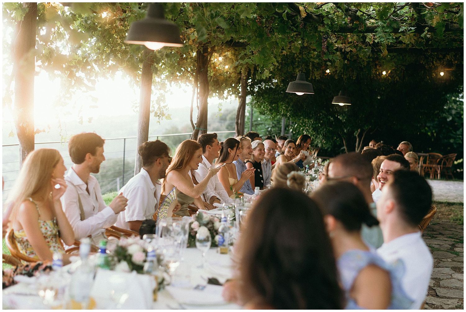 Wedding guests seated at a long outdoor dining table under string lights and trees with late afternoon sunlight.