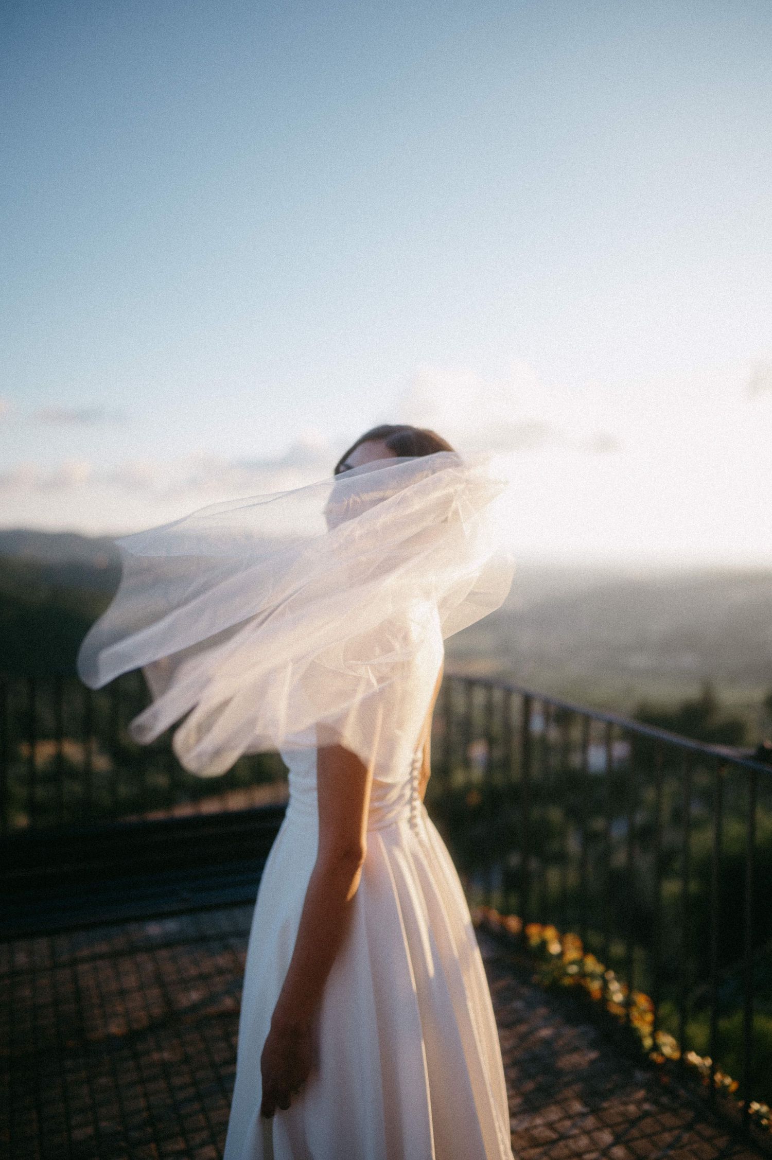 A flowing white wedding dress and veil catch the wind against a scenic hilltop vista at sunset.