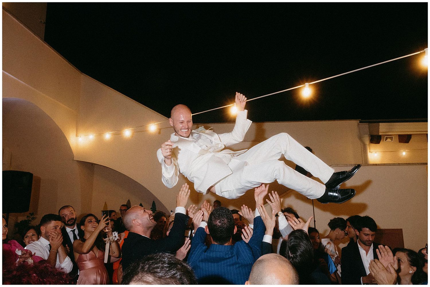 Wedding guests lift and toss someone in all white attire during an evening celebration with string lights overhead.