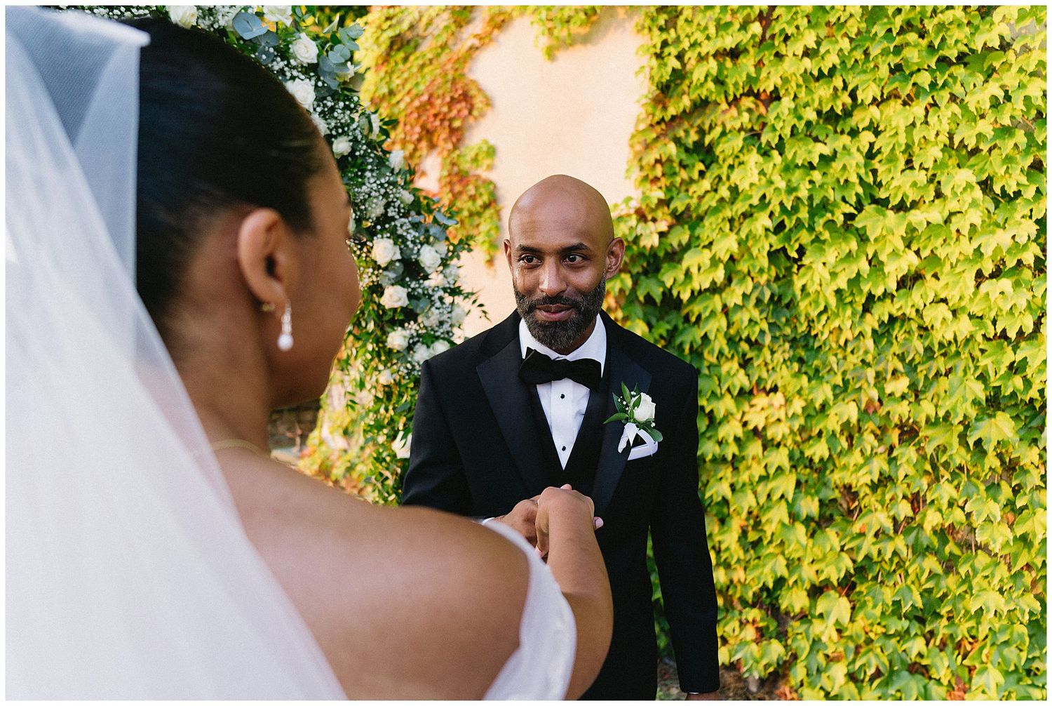 A wedding ceremony moment with groom in black tuxedo and bowtie against an ivy-covered wall under natural sunlight.