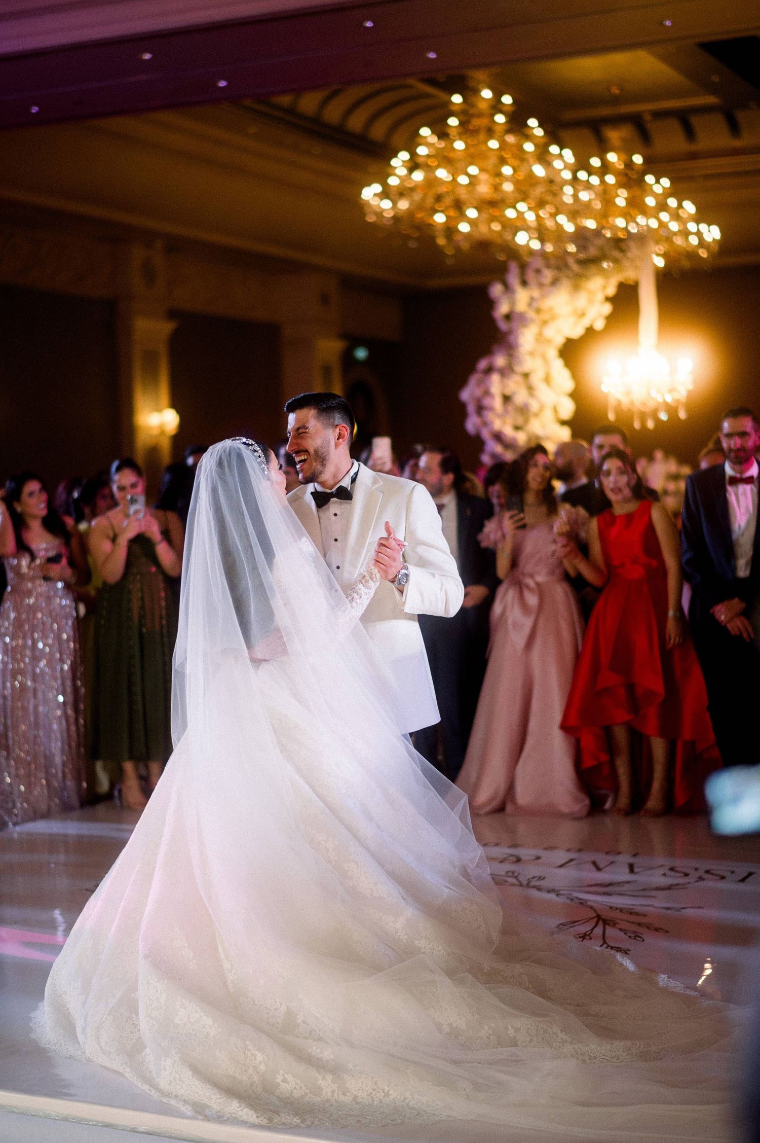 A couple shares their first dance at a wedding reception under a stunning chandelier while guests watch in formal attire.