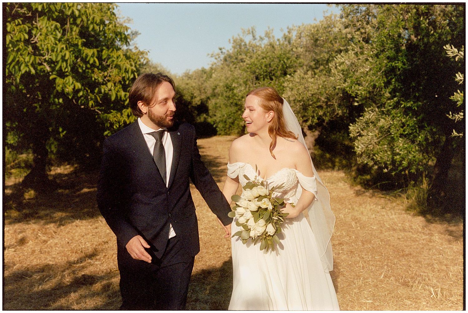 A couple walks on a dirt path surrounded by greenery during an outdoor wedding ceremony on a sunny day.