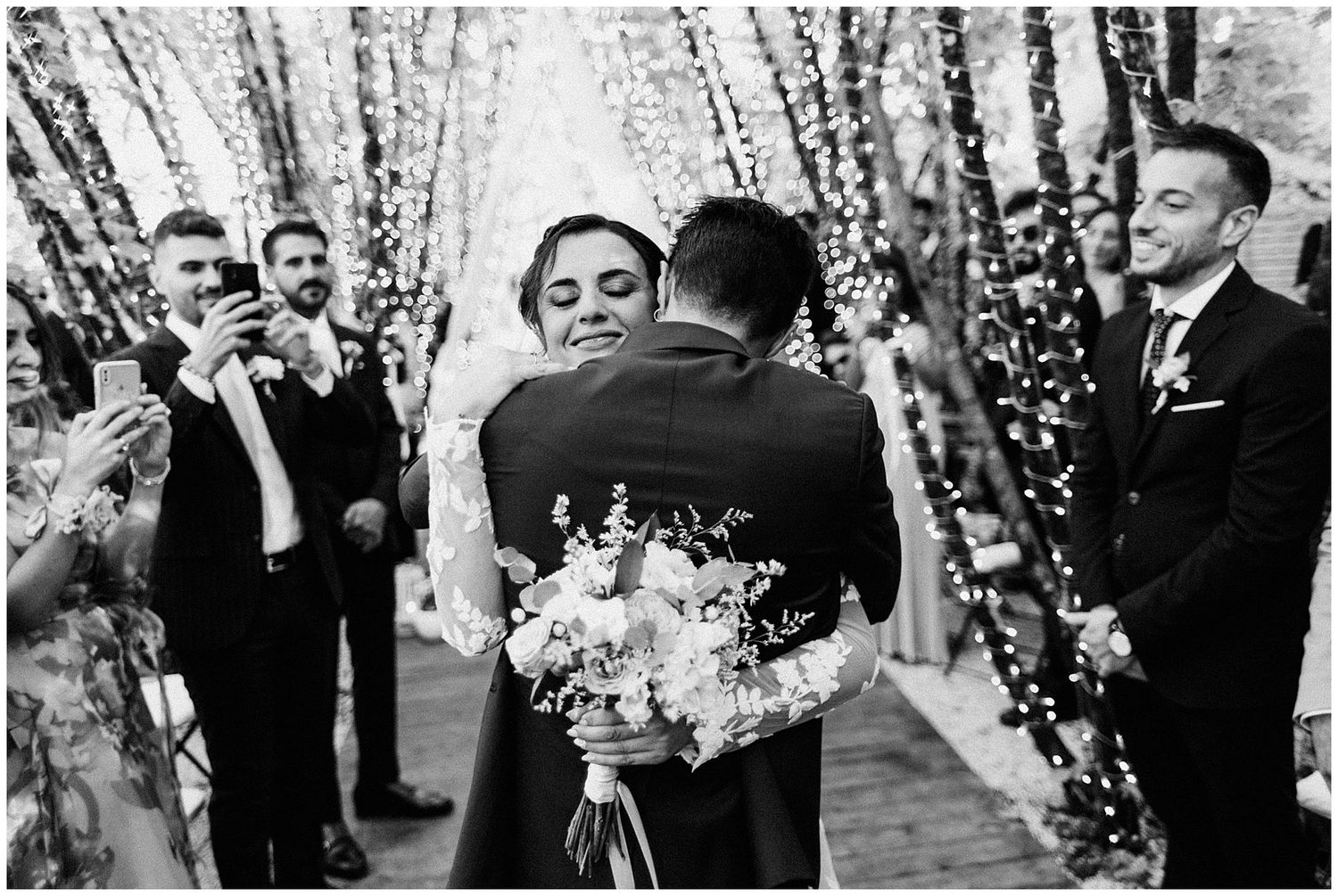 Wedding ceremony in progress under illuminated birch trees with gathered guests watching.