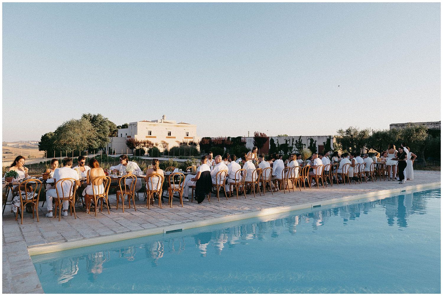 Elegant poolside wedding reception with guests seated at long tables beside a blue swimming pool.