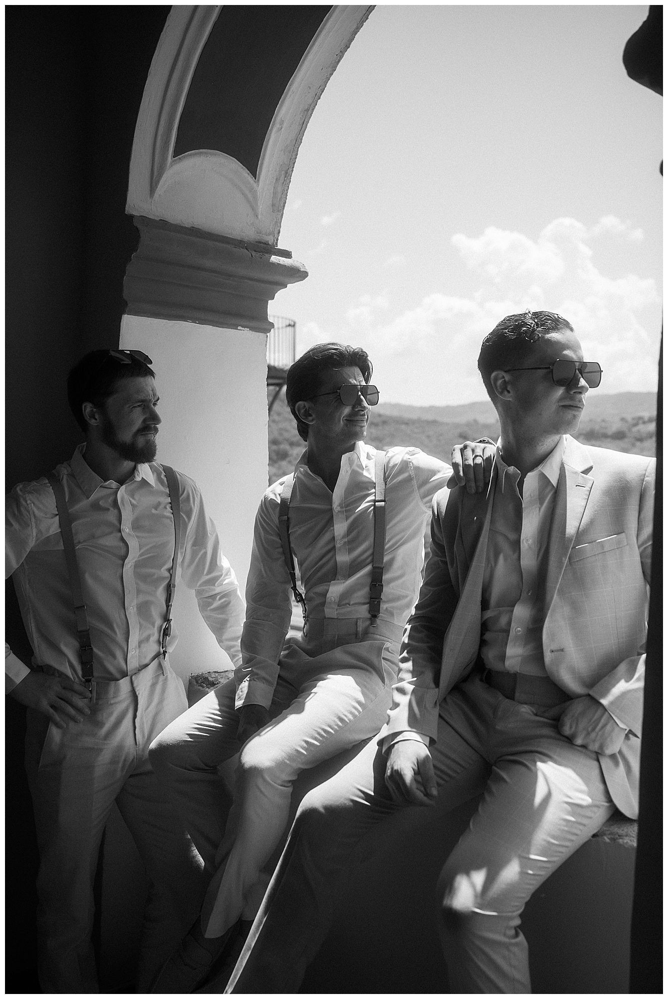 Three people in light-colored suits sit together on a balcony in a black and white vintage-style photograph.