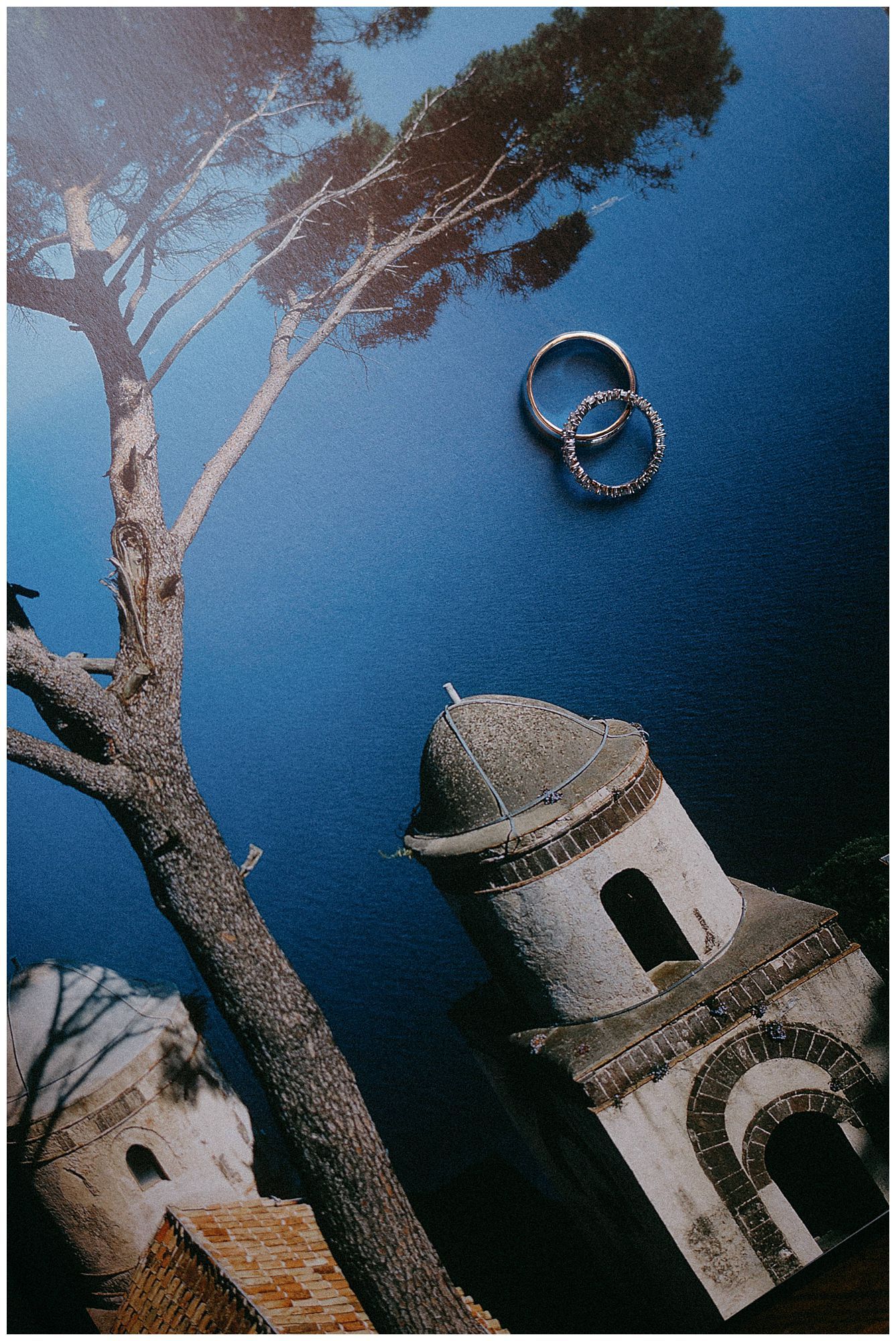 Wedding rings float above a vintage church bell tower and tree branch against a moody blue evening sky.