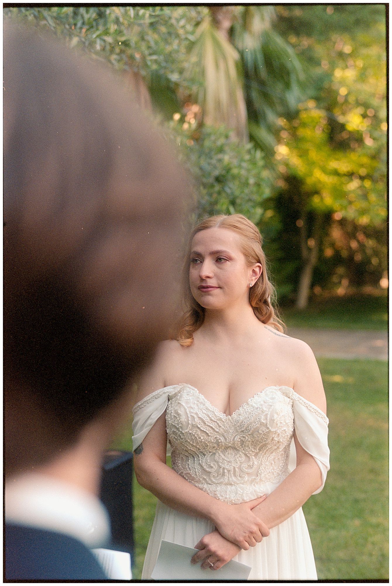 A person wearing an elegant off-shoulder white lace wedding gown stands in a sunlit garden setting.
