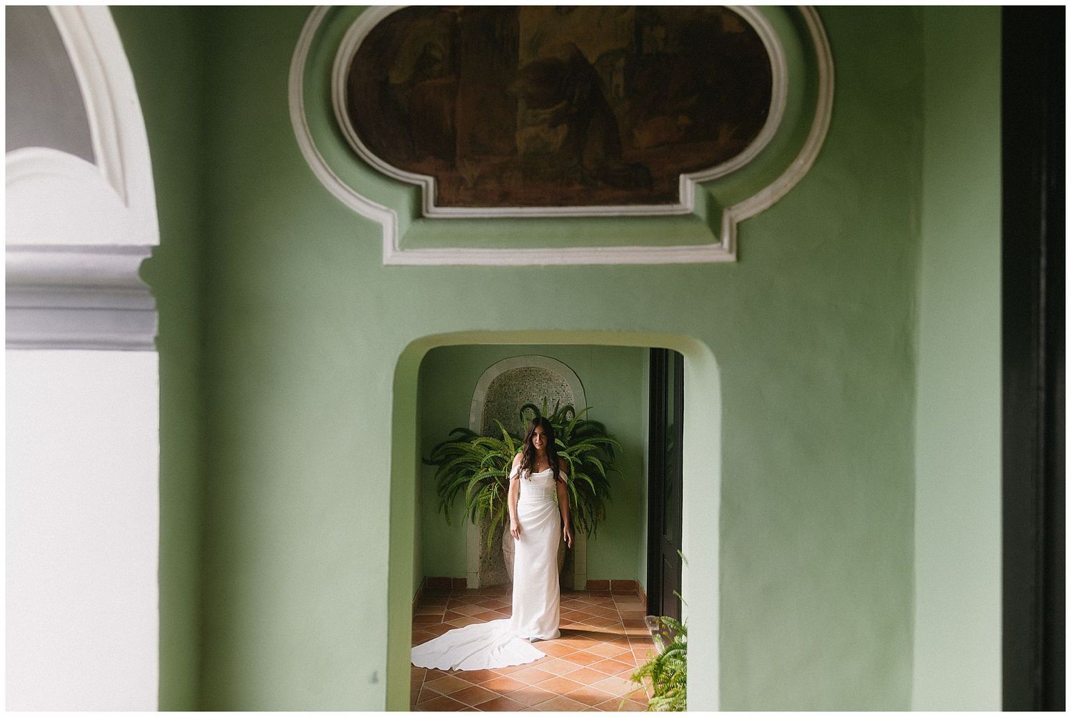 A person in white stands in a pale mint green archway with ornate architectural details and indoor plants.