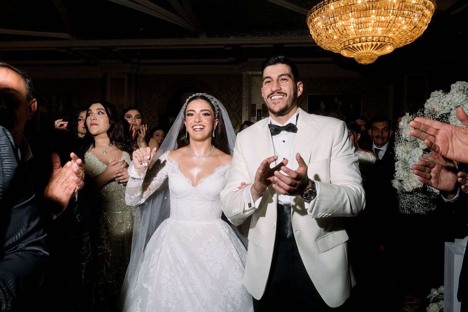 A joyful couple celebrates during their wedding reception under a crystal chandelier while guests applaud.