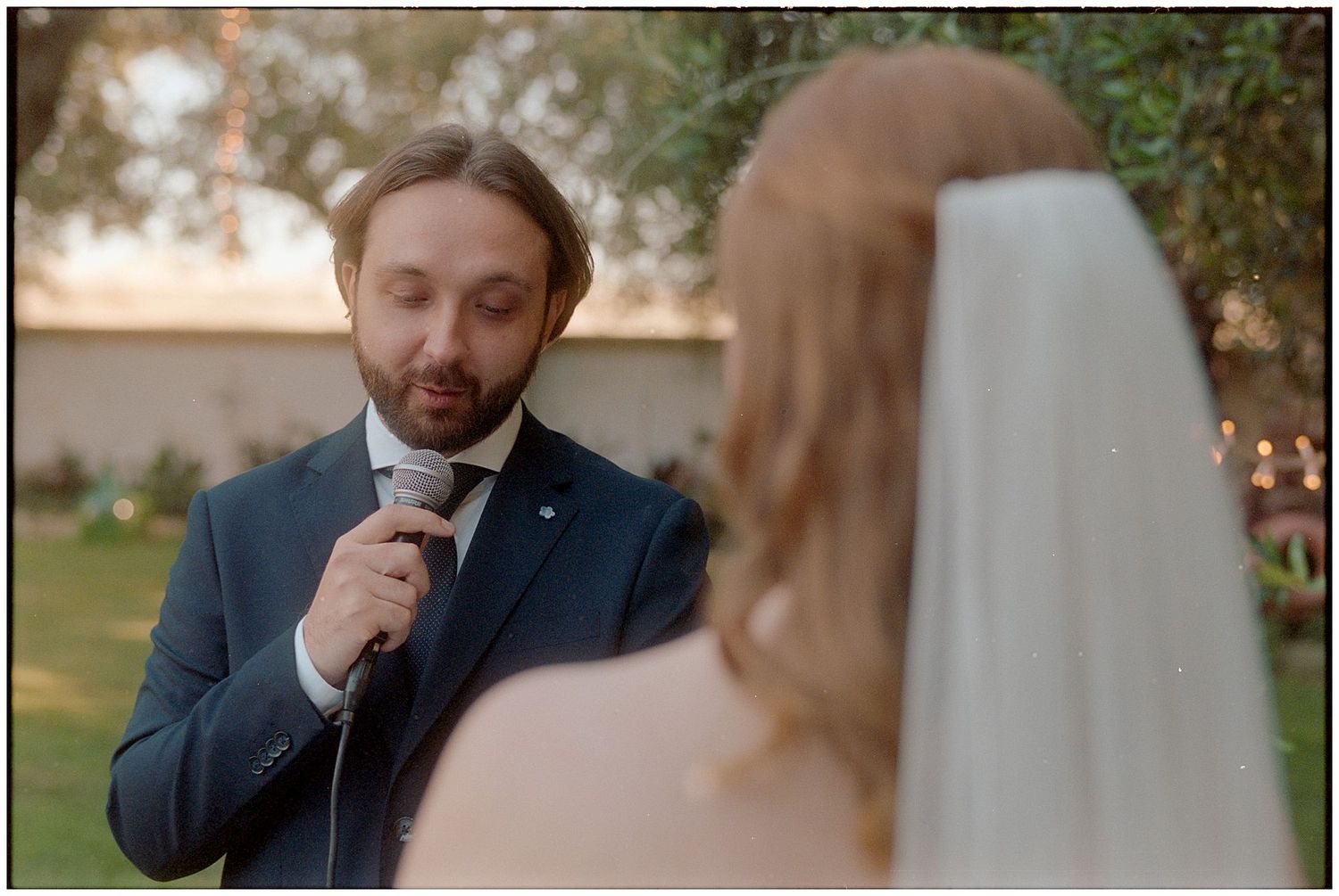 Groom in navy suit speaks into microphone during outdoor wedding ceremony with string lights in background.