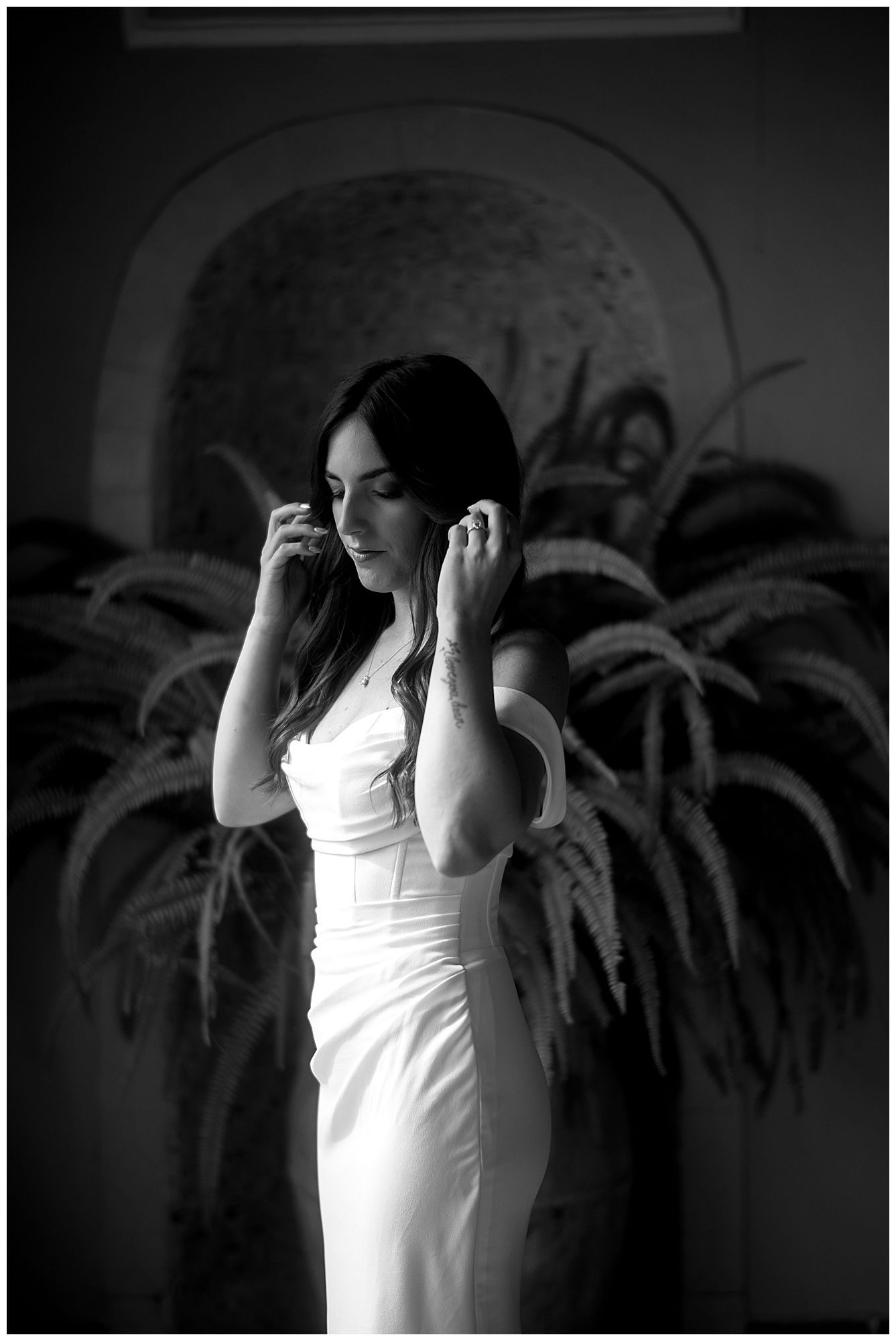 Black and white photograph of someone in an elegant white dress posing against a wall with palm tree silhouettes.