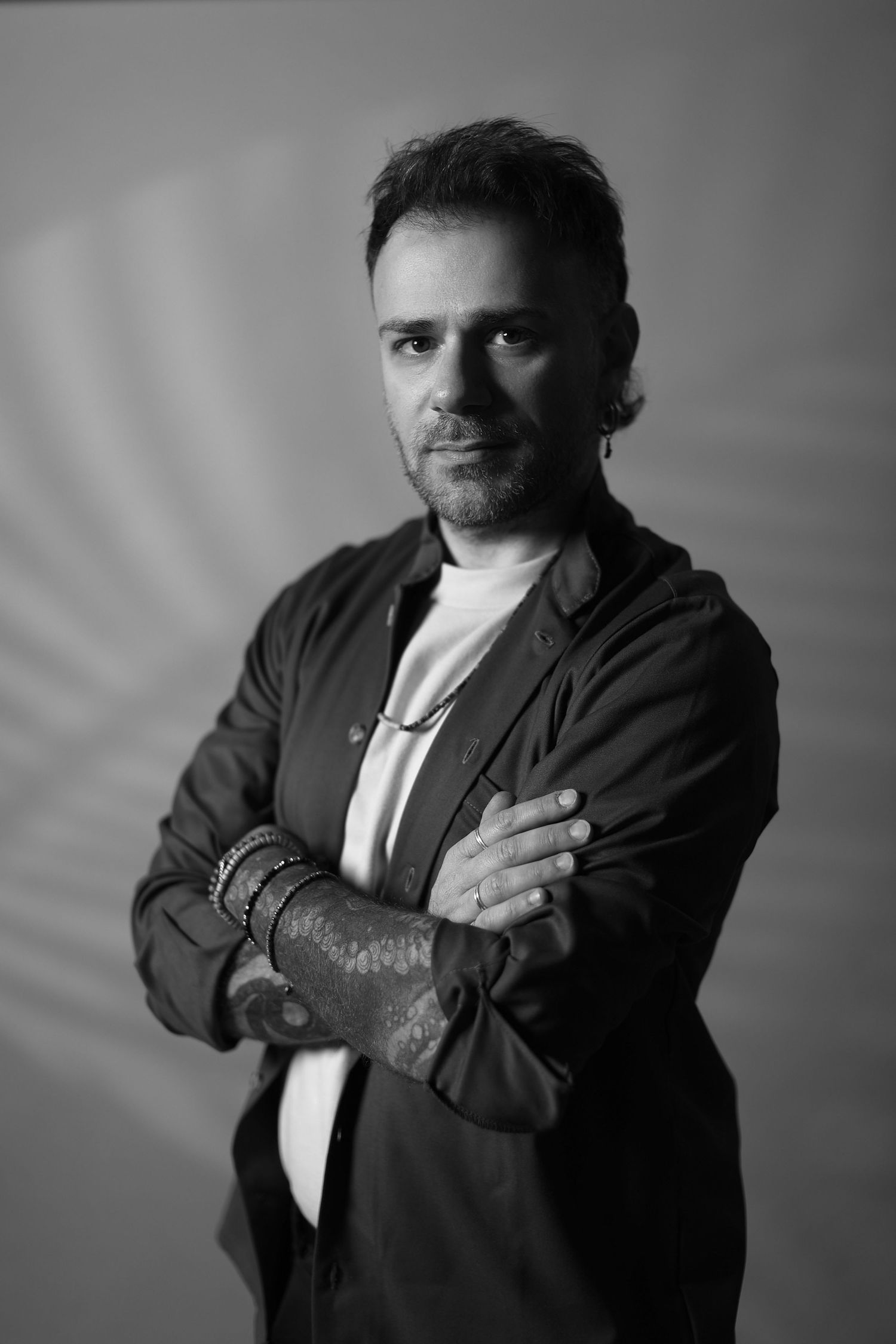 Black and white portrait of person in casual jacket with arms crossed and tattoos visible against neutral backdrop.
