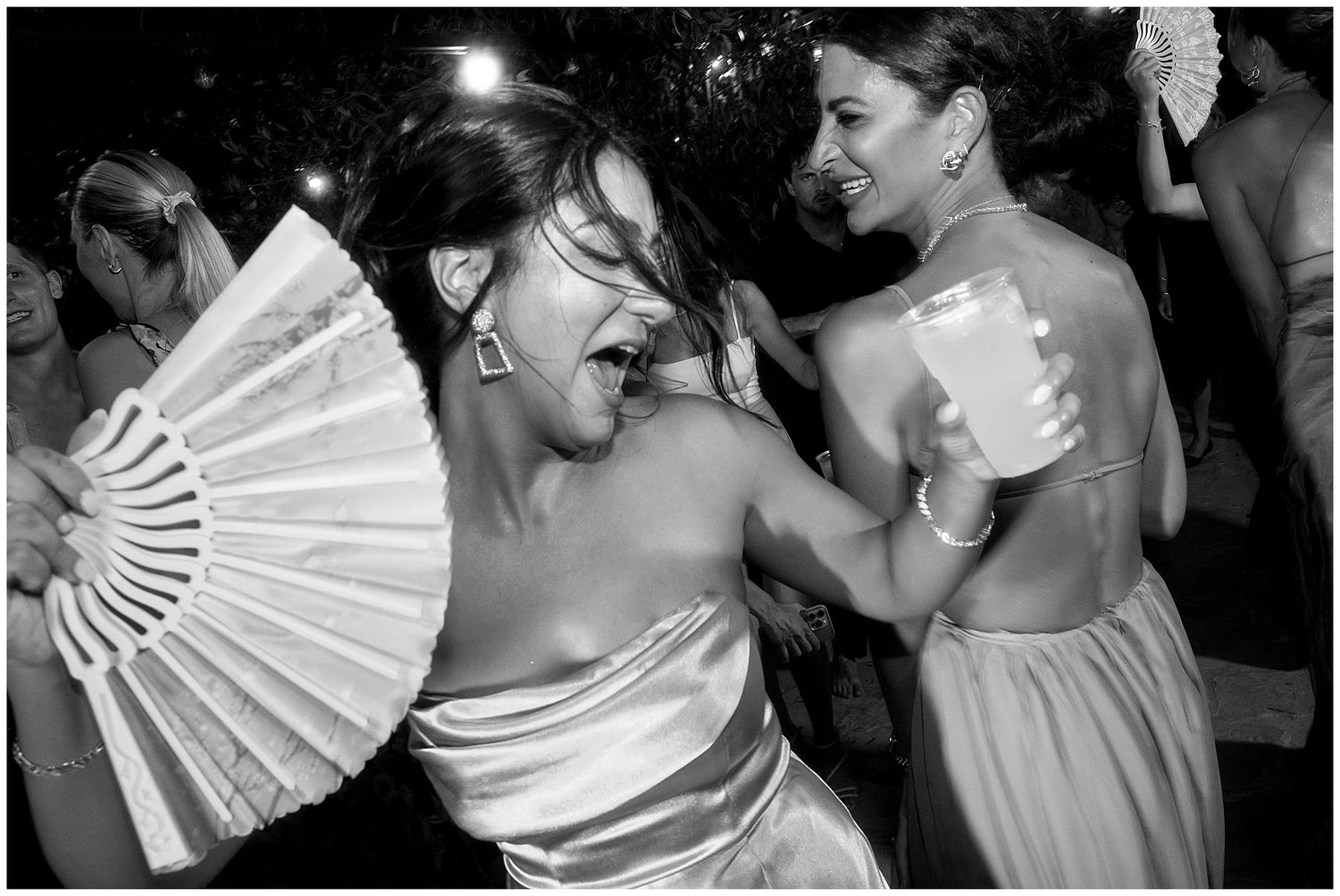 Two wedding guests dancing enthusiastically at a reception, one holding a white paper fan and both wearing light dresses.