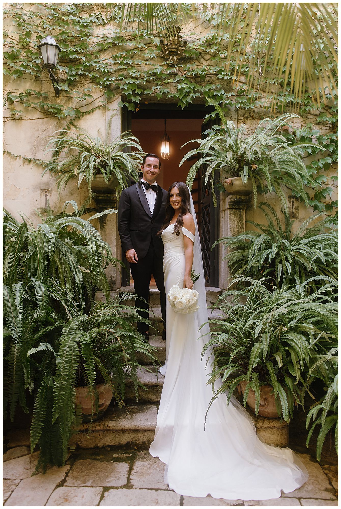 Couple standing on garden steps of an elegant venue surrounded by lush ferns and greenery.