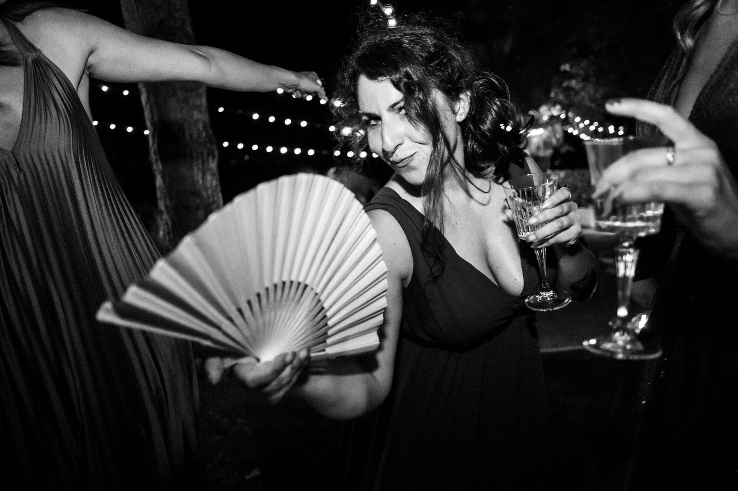 A person in dark attire holds a decorative fan while celebrating at a party with string lights.
