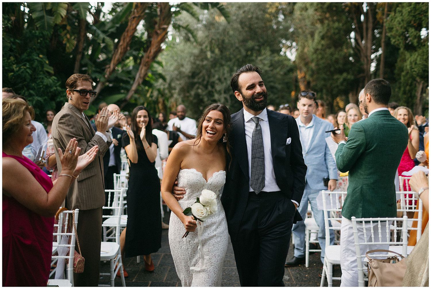 A joyful couple walks down an aisle surrounded by guests at an outdoor garden wedding ceremony.