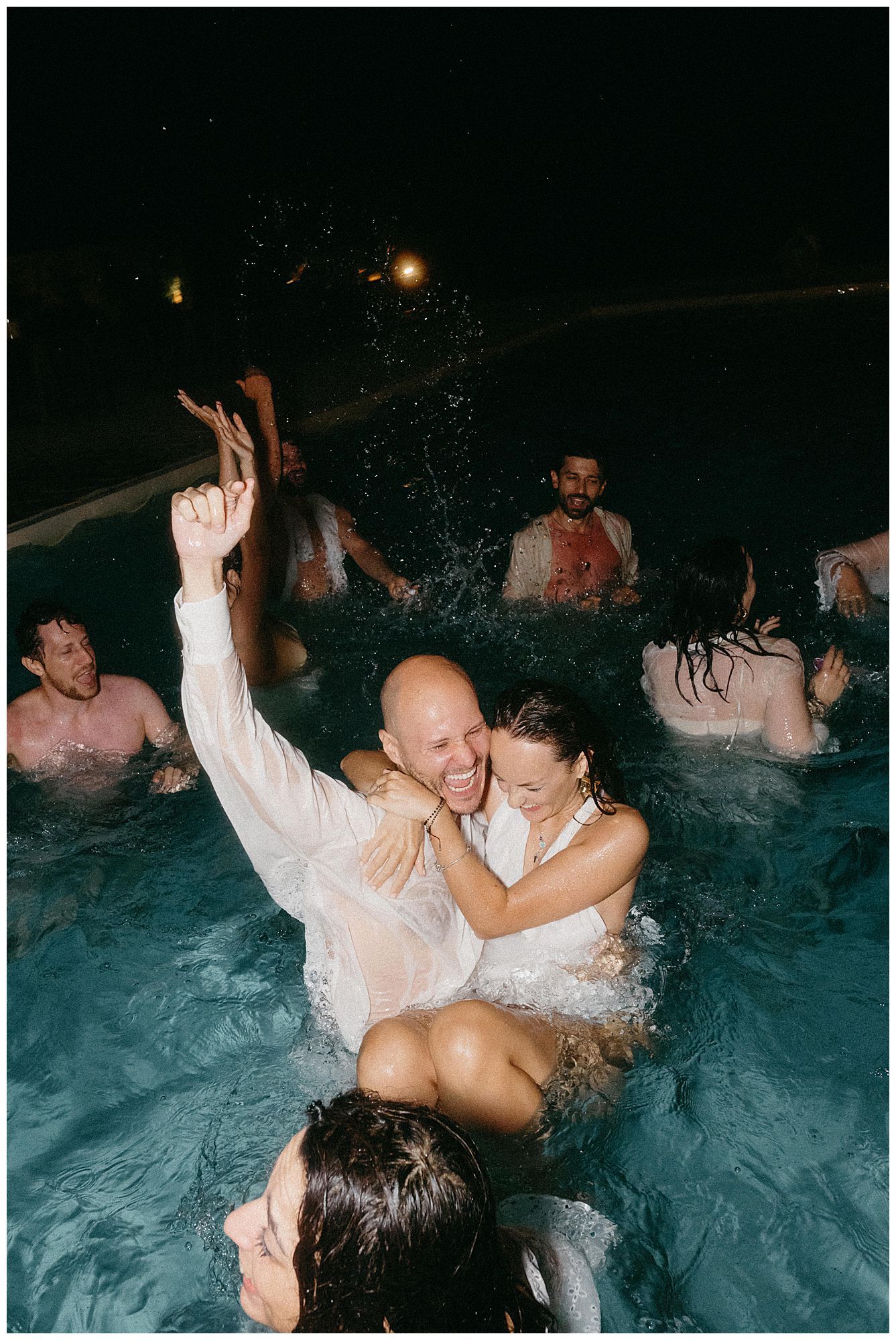 Happy people celebrating and splashing in a pool at night with festive lighting in the background.